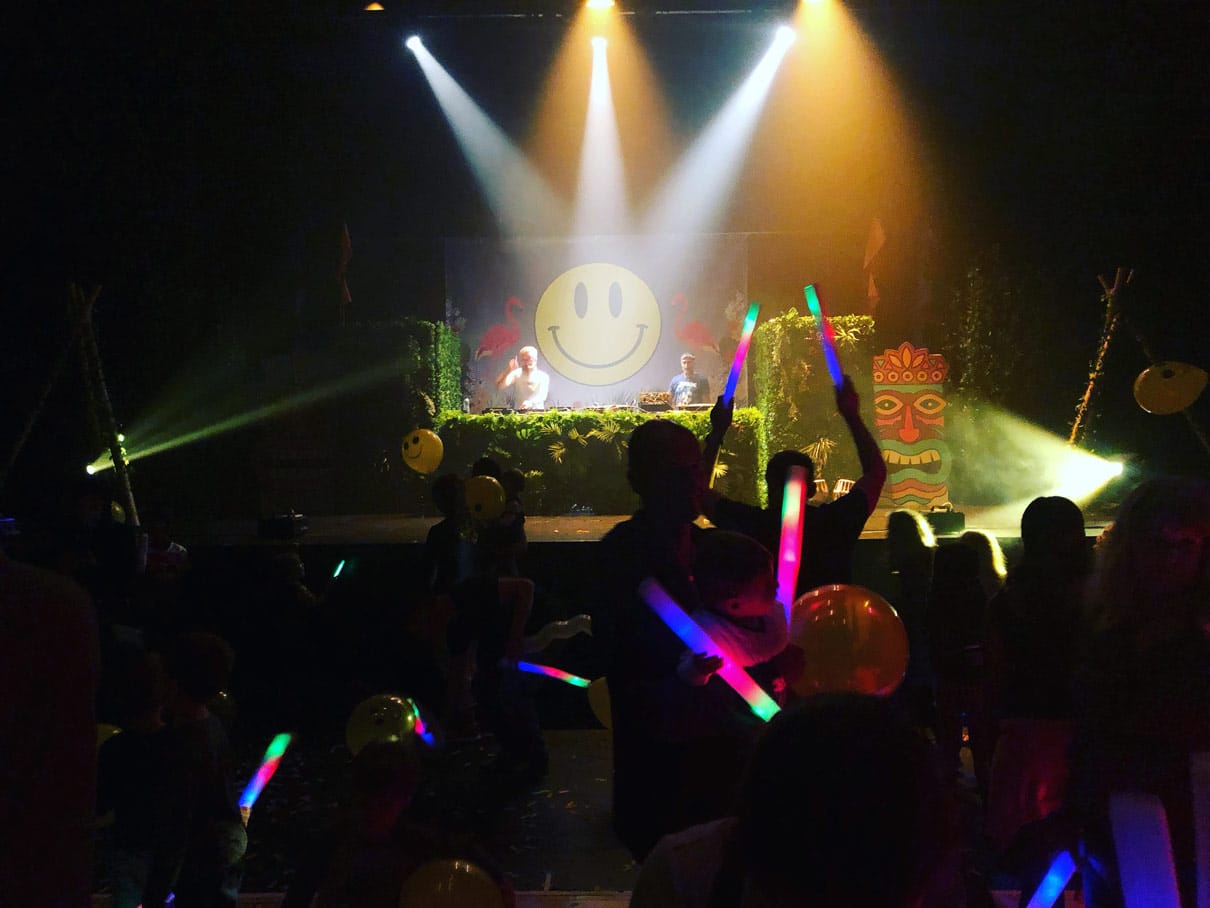 On the Pentland Theatre stage are two DJs behind decks covered in fake grass and greenery, behind them is a projection of a large smiley face and flamingos. In the foreground, a crown of people are dancing holding glow sticks.