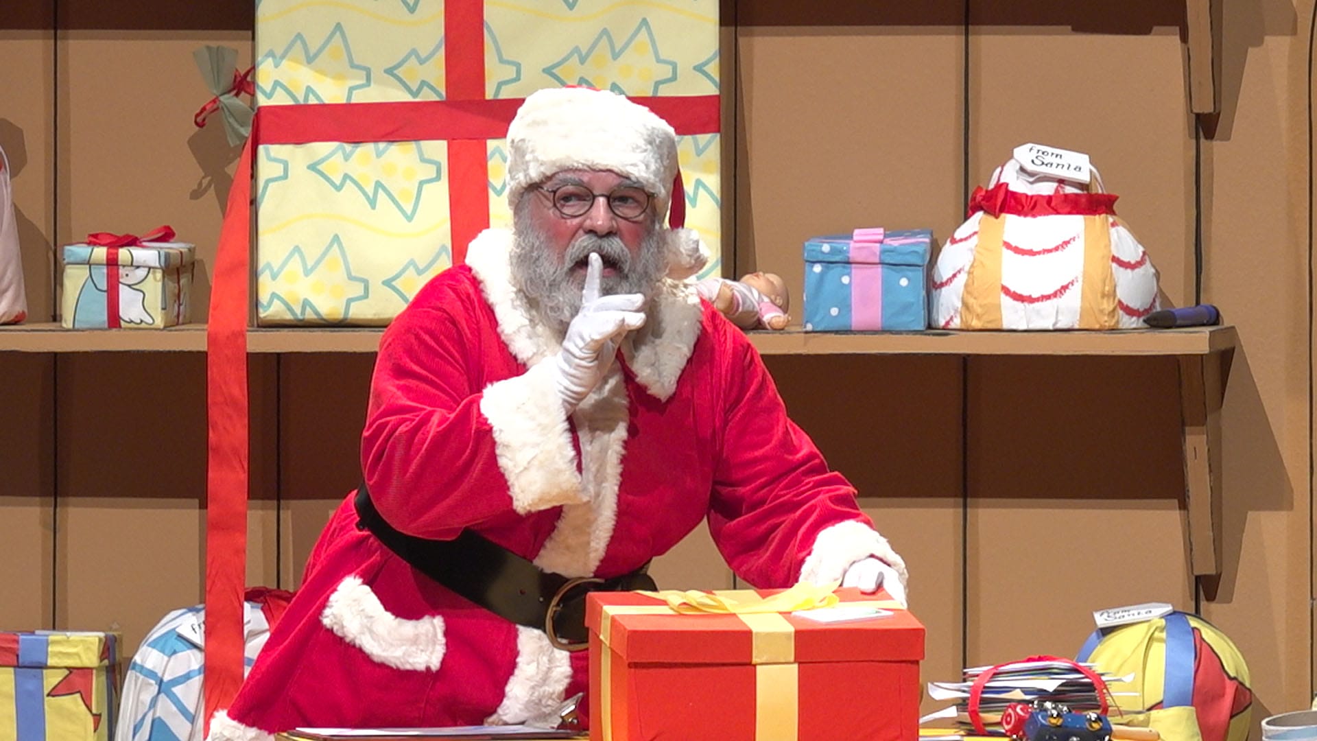 Santa does a 'shhh' sign with his hands while holding a present