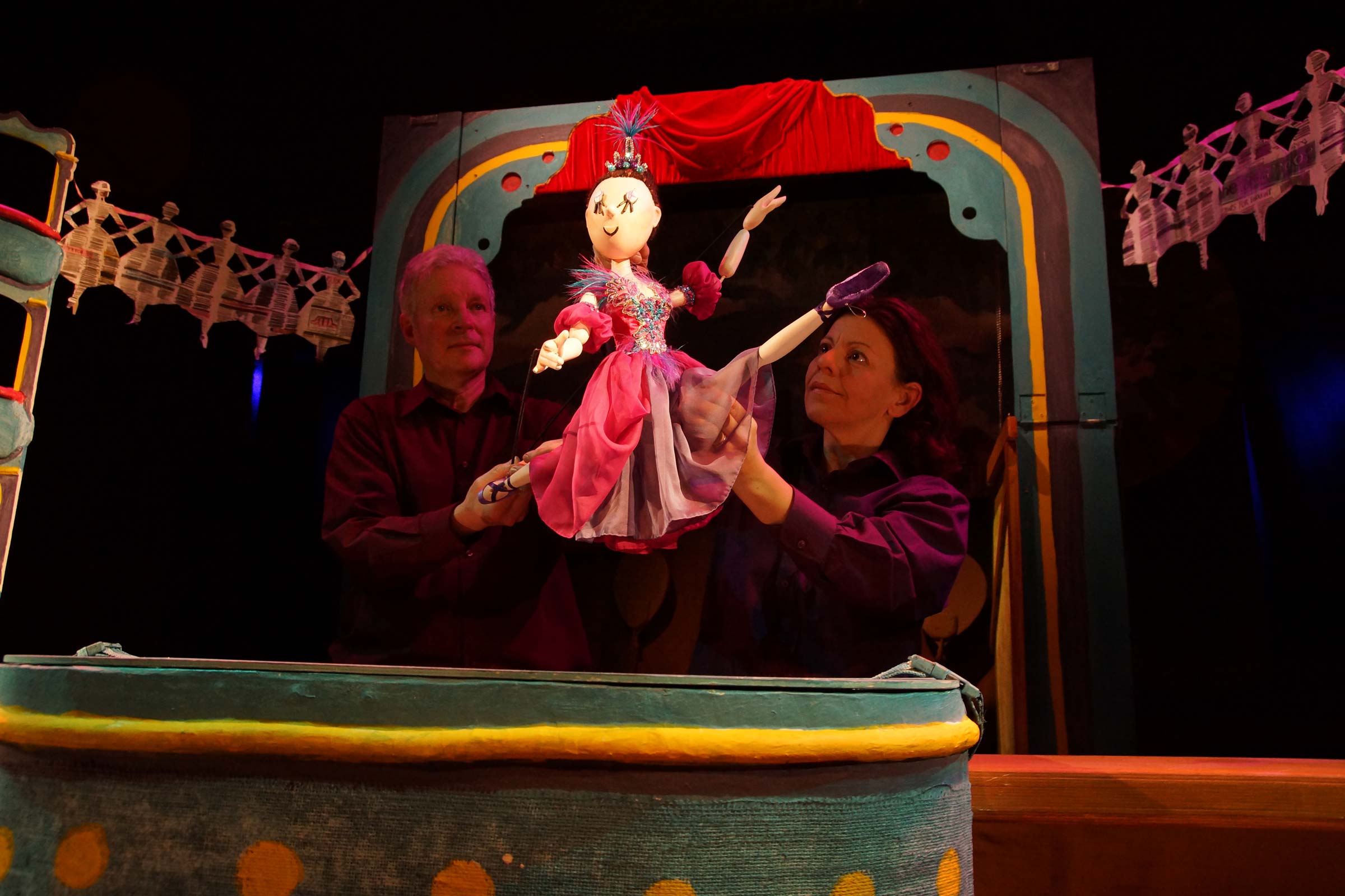 Two puppeteers hold a wooden ballerina puppet as she leaps through the air