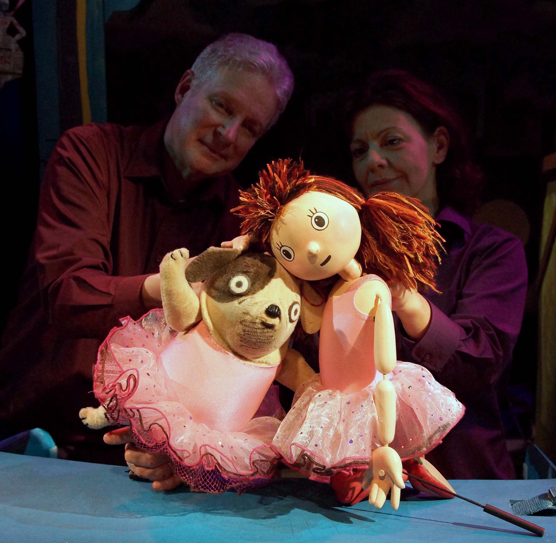 Two puppeteers hold puppets of Biff, a small dog wearing a pink ballet outfit, and Anna, dressed the same. Anna and Biff are sitting close together with their heads touching.