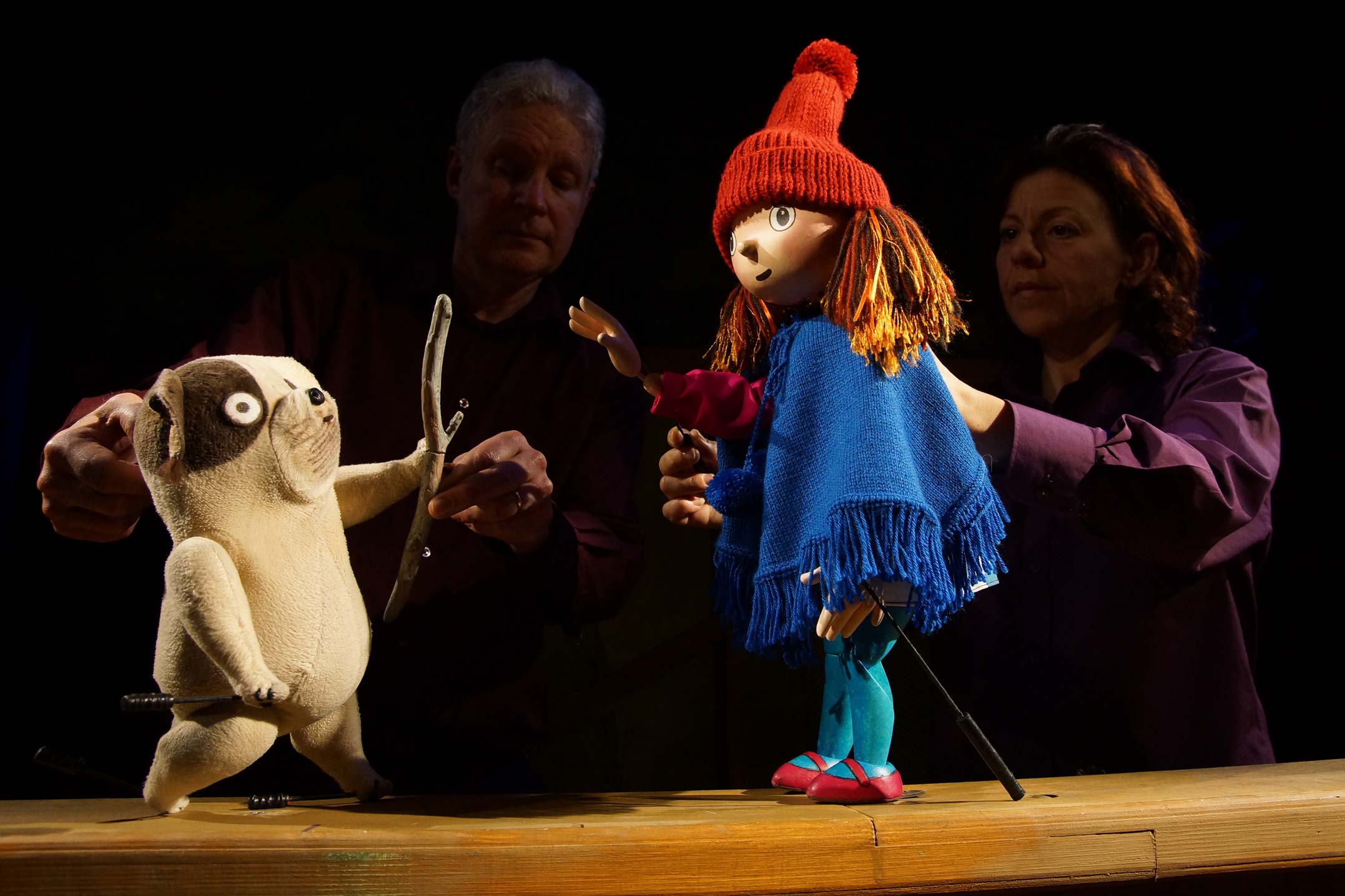 Two puppeteers hold puppets of Biff, a small dog, and Anna, a young girl wearing an orange hat and a blue knitted poncho.