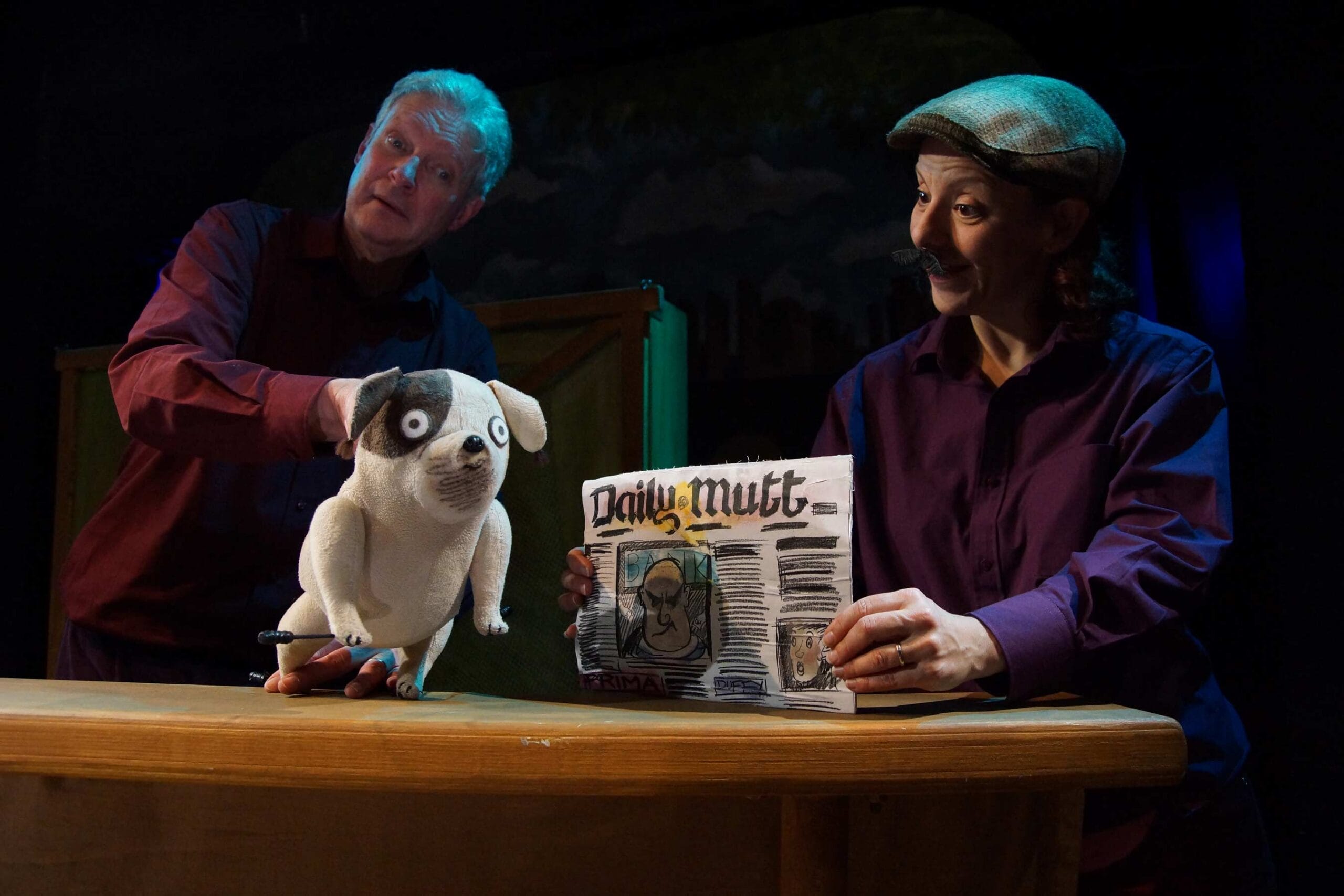 A puppeteer holds a puppet of Biff, a small dog, whilst an actor dressed as a news-seller holds a copy of a newspaper called The Daily Mutt