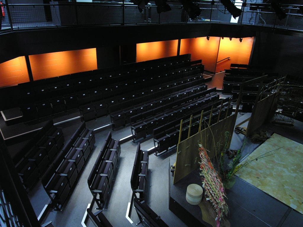 The Studio Theatre, pictured from above. The stage is set for a performance with screens and a light green mat on the stage. The room has rows of black upholstered seats and an orange wall at the back.