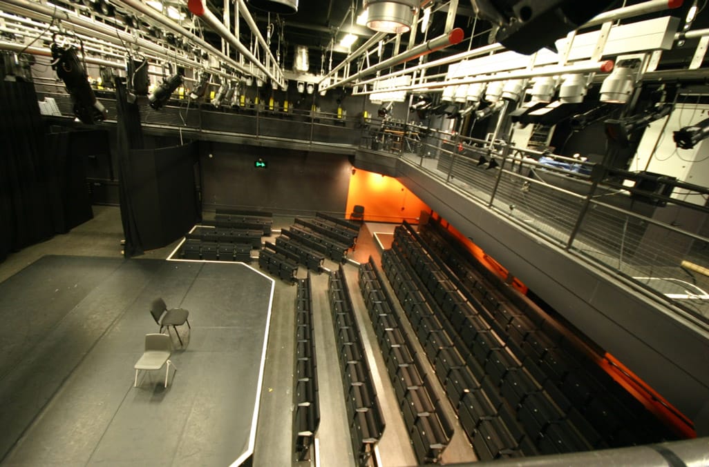 The Studio Theatre pictured from above. A black covered thrust stage and rows of black upholstered seating can be seen. Silver lighting bars with stage lights attached can be seen at the top of the image.