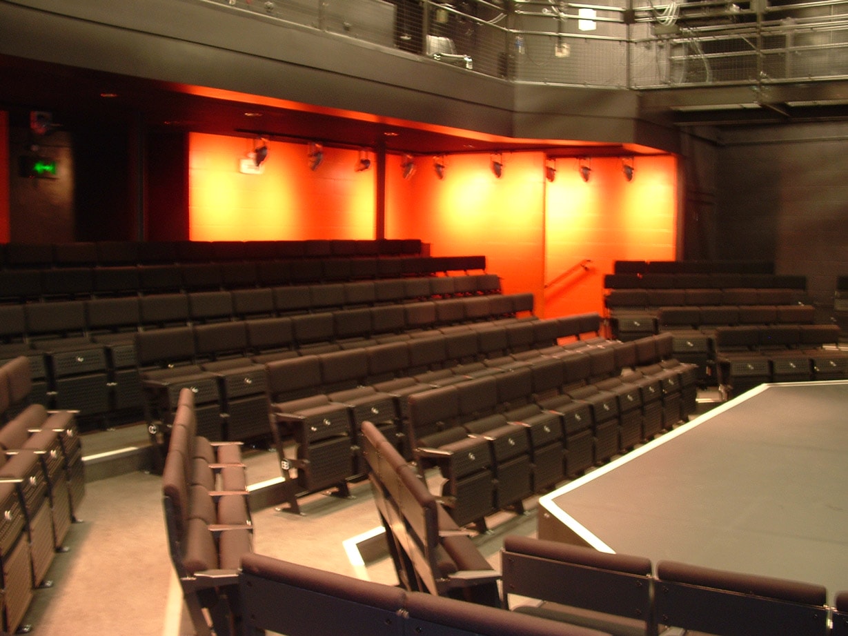 The Studio Theatre, pictured from the seats at the side of the stage. The front of the thrust stage can be seen with rows of black upholstered seating wrapping around it. An orange painted wall can be seen at the back of the room.