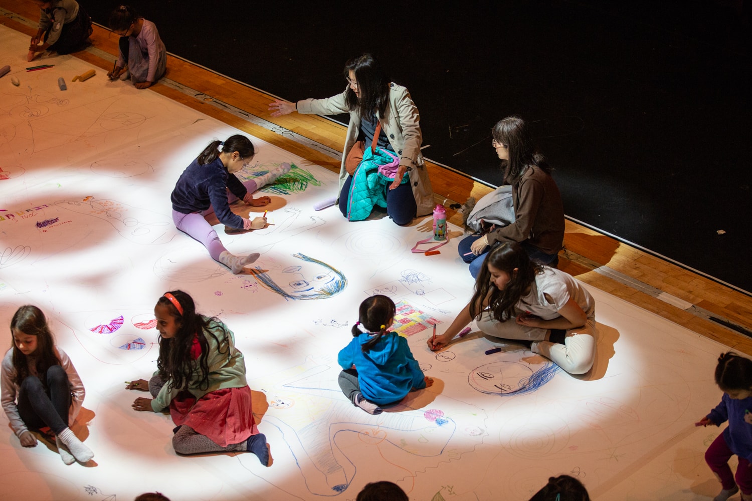 An interactive community art project. A large canvas spreads across the floor where families and young people are drawing and making art. Projected lights illuminate the canvas in abstract shapes.