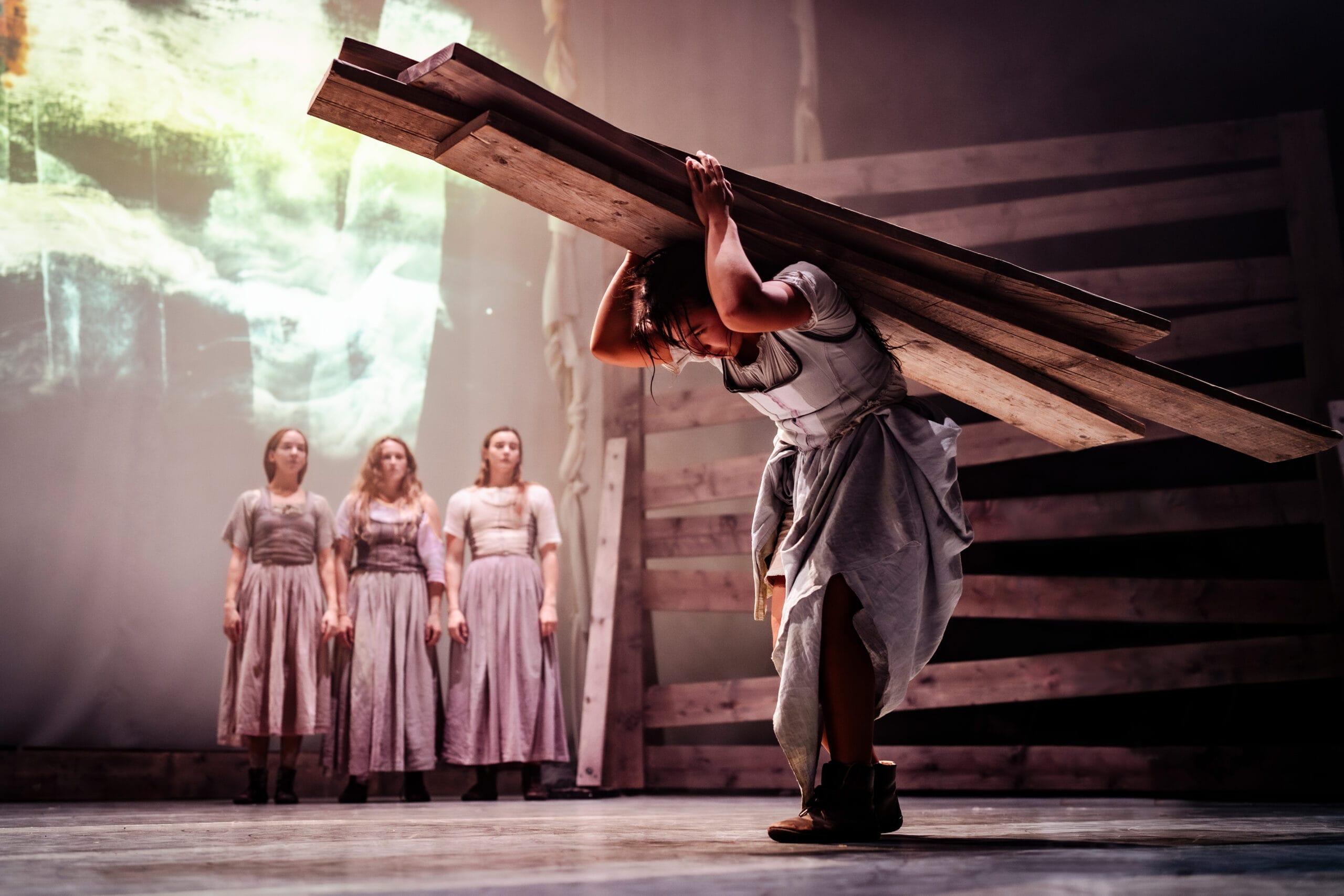Tess performer holds heavy planks of wood above her head, as female ensemble watch from the background. The image has a lilac tint with deep shadows across Tess as she heaves the weight of the wood on her back.