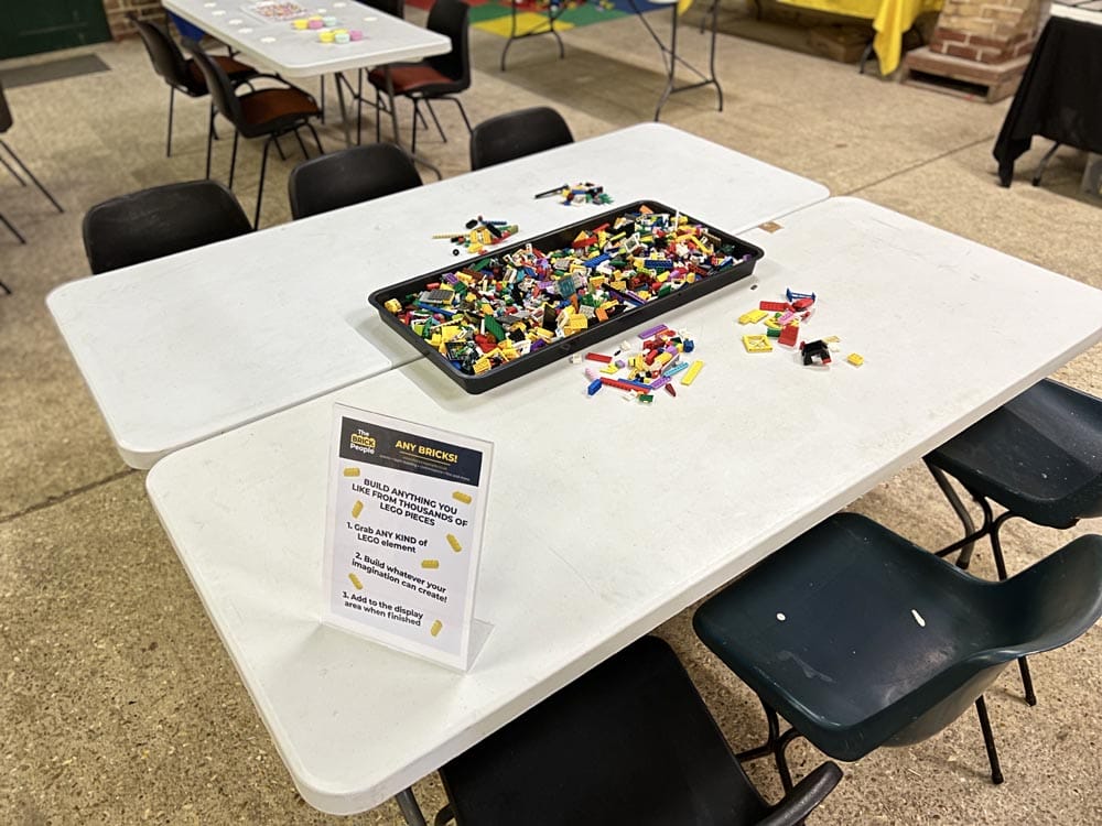 Tables and chairs are set up for a lego building activity. In the centre of the table is a tray full of colourful lego pieces, with a sign on the table giving participants ideas for their builds.