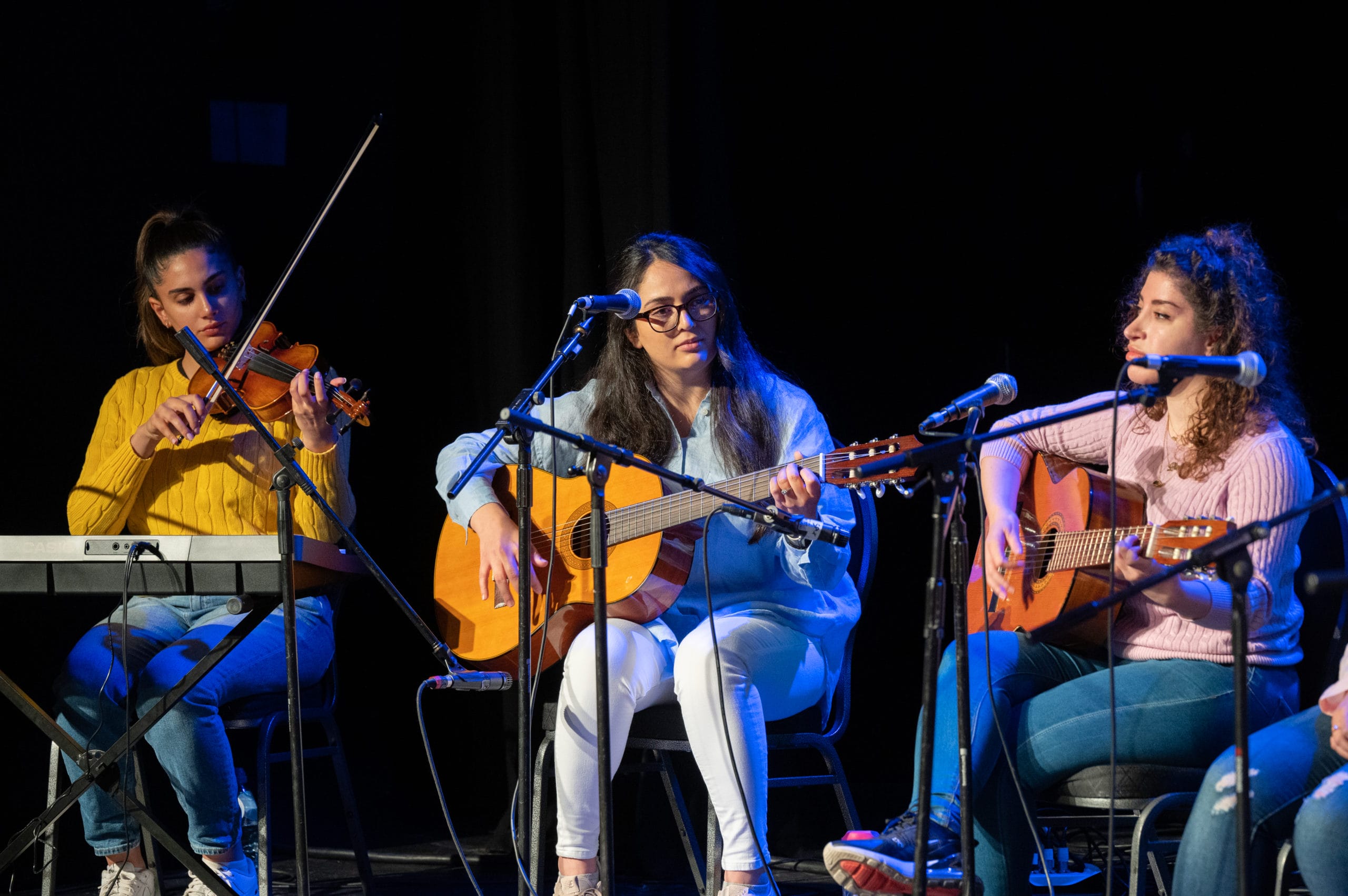 Three female musicians onstage. A violinist on the left and two acoustic guitarists on the right. They are all wearing jeans and sweatshirts.