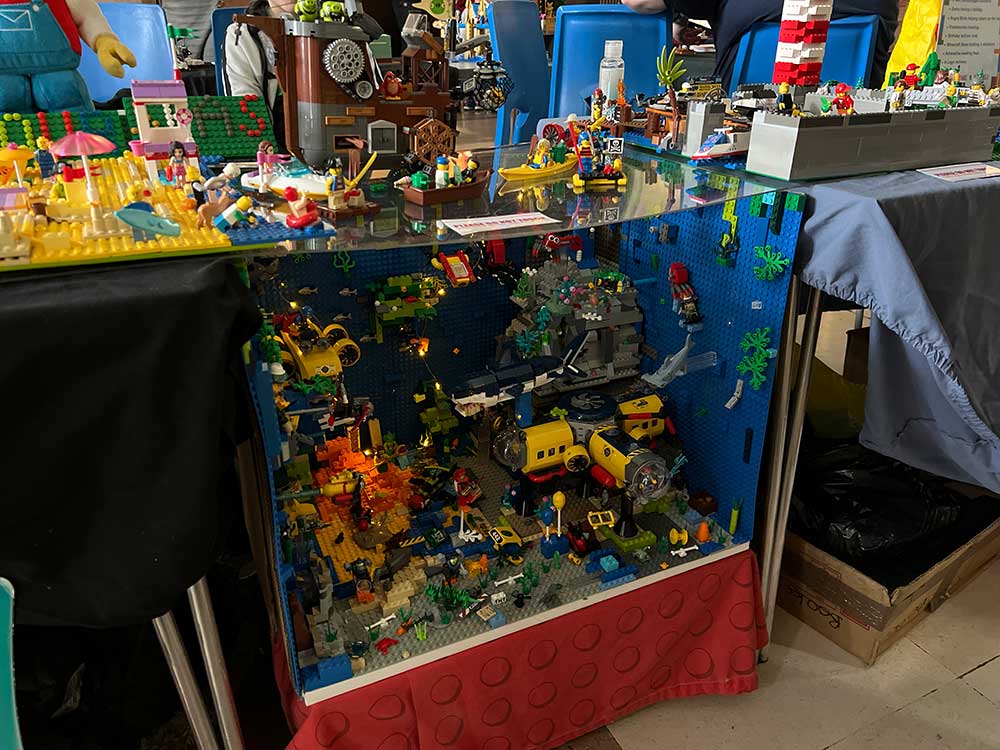 A glass cabinet is packed full of intricate lego builds. Vehicles, houses, plants, even mermaids on the beach.