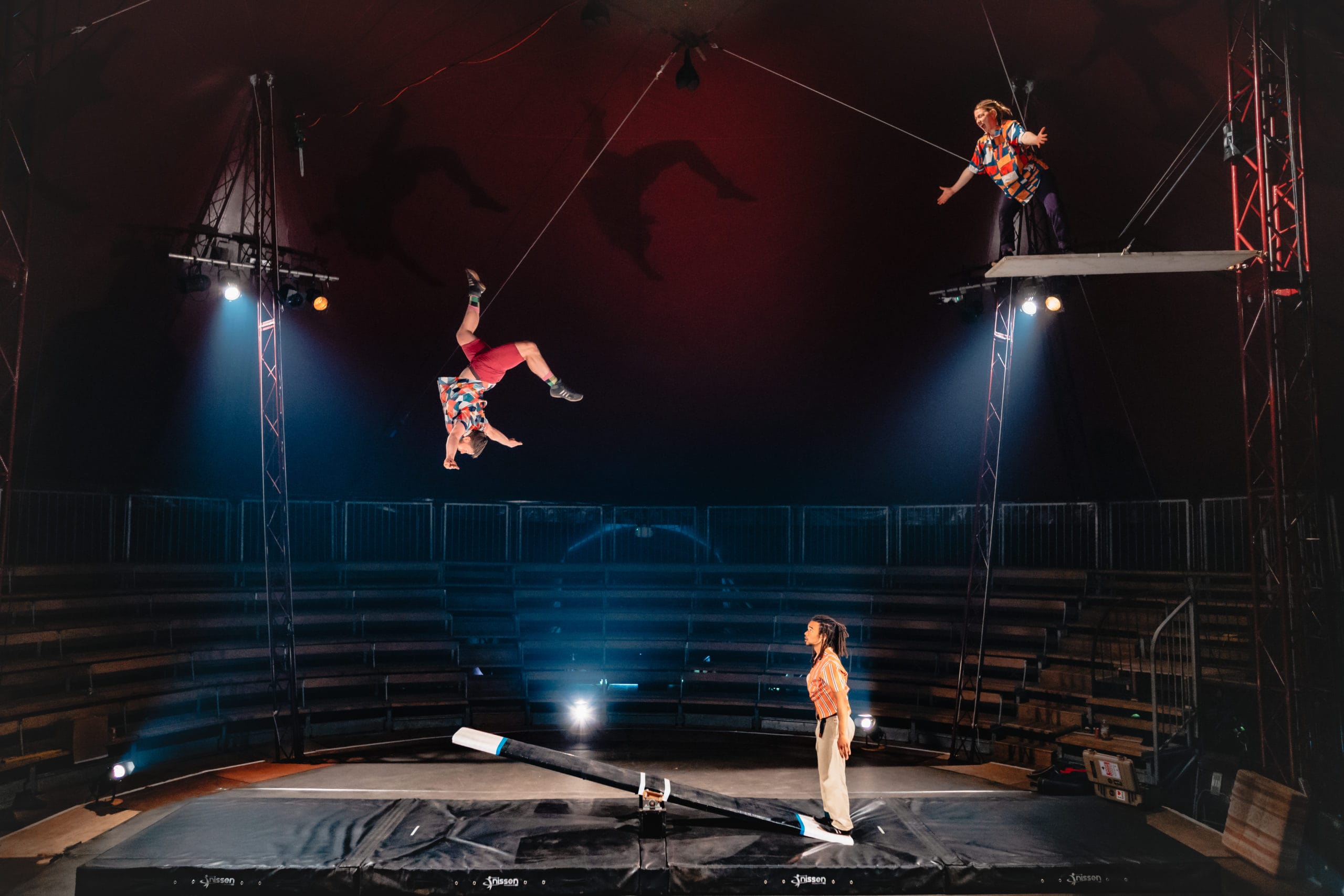 Three Revel Puck circus performers engage in an impressive stunt. One performer stands on the end of a small seesaw, whilst another is catapulted high into the air, and is performing a backflip upside down. A third performer stands on a plank suspended high in the air, ready to catch their fellow performer.