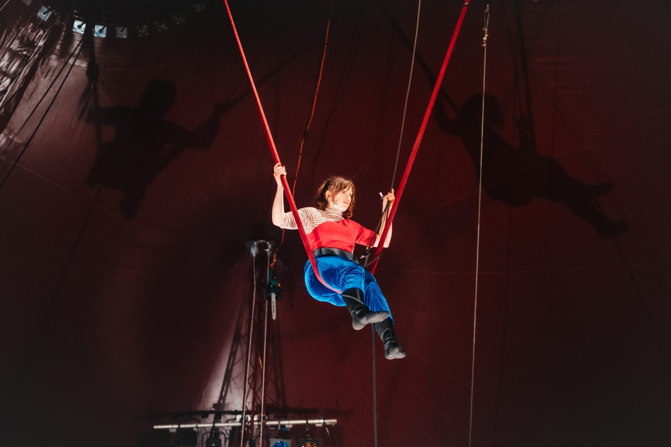 A Revel Puck circus performer is sat in a swing-like position on a high trapeze. The trapeze rope is red and the performer wears a red shirt and vibrant blue trousers.