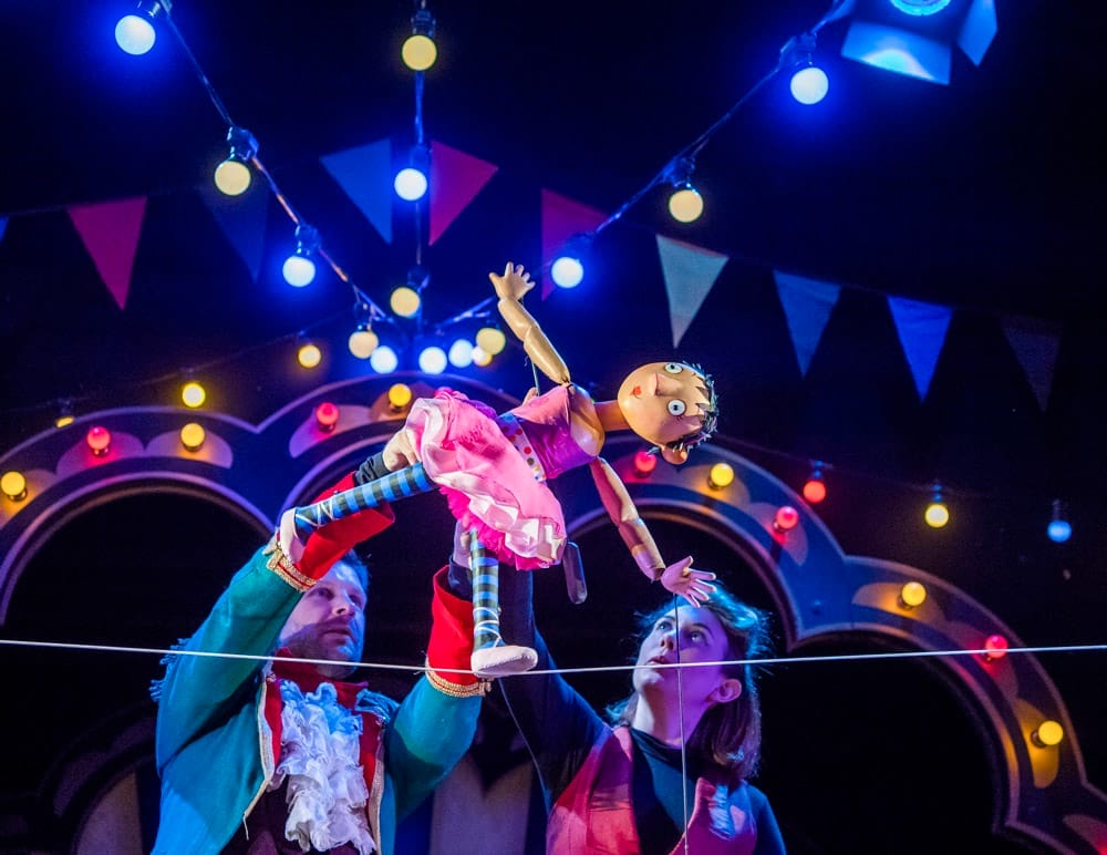 Two puppeteers hold a puppet of a circus performer as she walks the tightrope. Behind them can be seen the lights of a circus tent and bunting