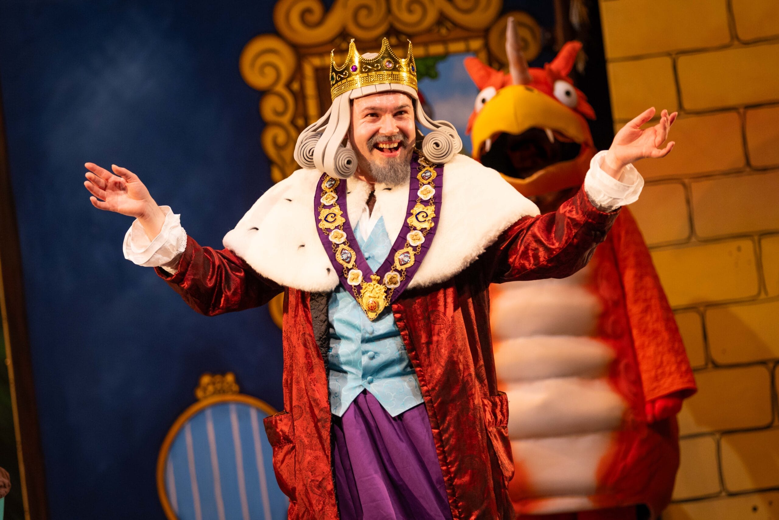 The King, in full regal regalia and with a long grey wig, holds his arms wide. Behind him can be seen Zog the orange dragon.