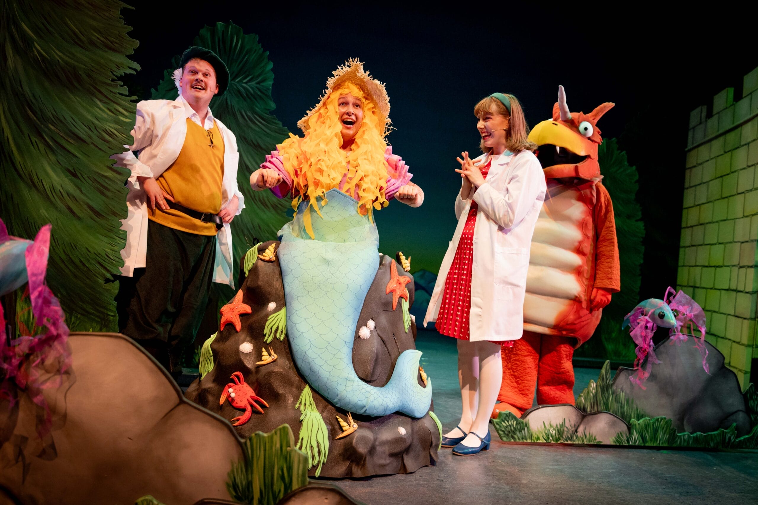 Princess Pearl and Sir Gadabout wearing lab coats stand either side of a mermaid who sits on a rock and has long blond hair and a green tail. Zog the orange dragon can be seen at the back of the stage.