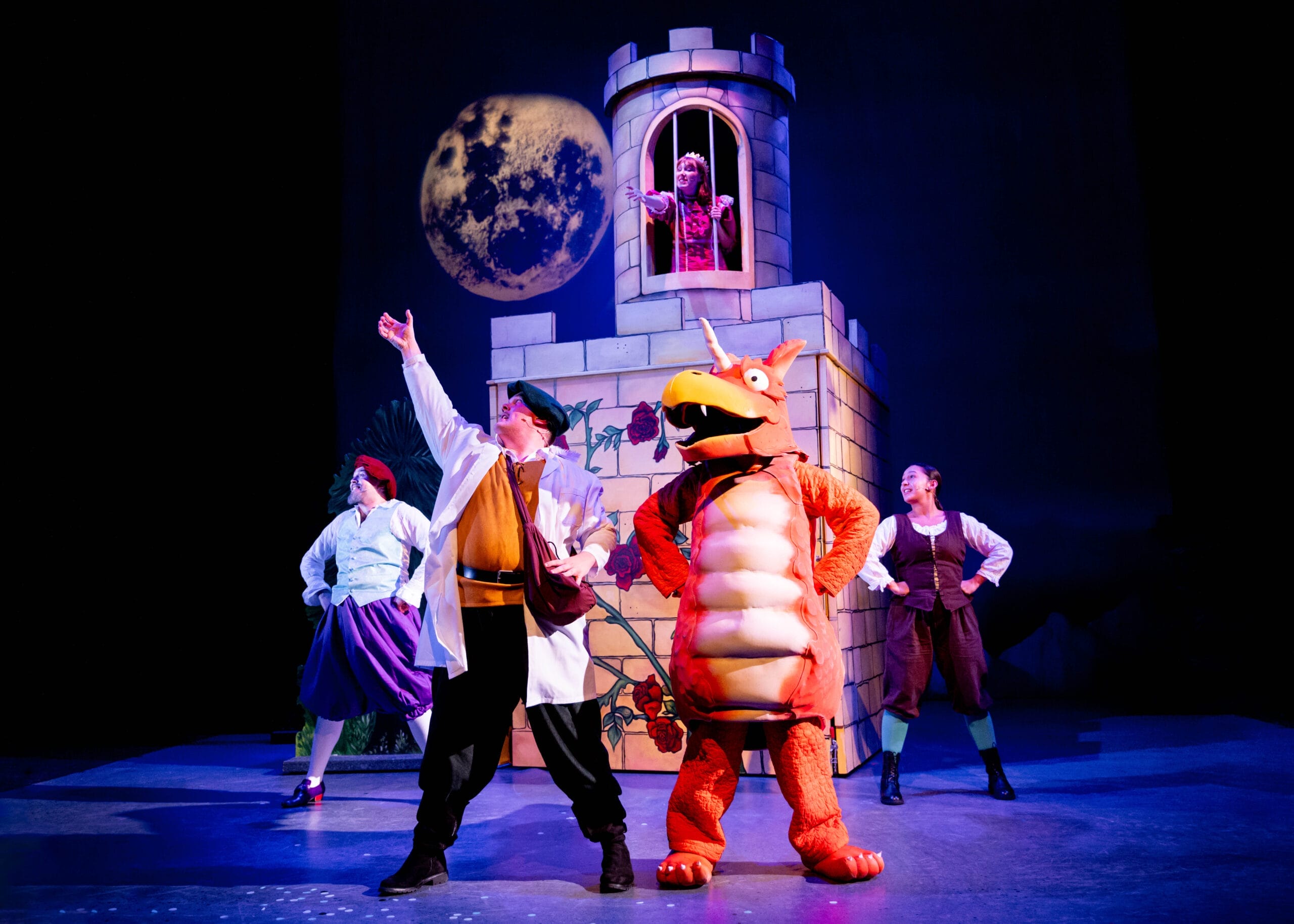 Zog the orange dragon stands at the front of the stage with his hands on his hips alongside 3 other performers in the same pose. Behind him is a castle in which the princess is trapped in a tower, she reaches her arms through the barred window.
