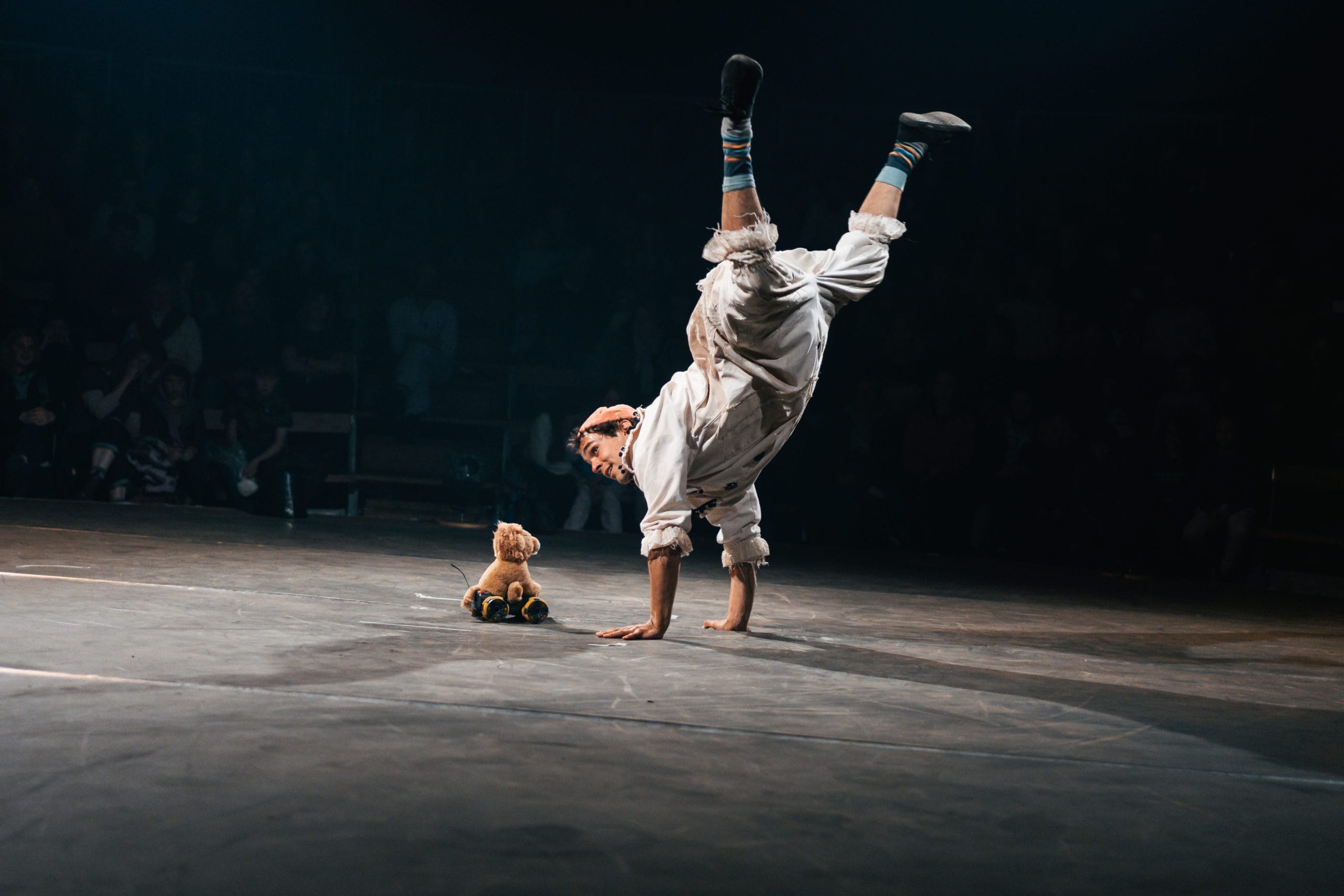 A performer engages in a handstand. They are wearing an oversized contemporary clown costume. On the ground next to them is a remote controlled robot, part of the act.