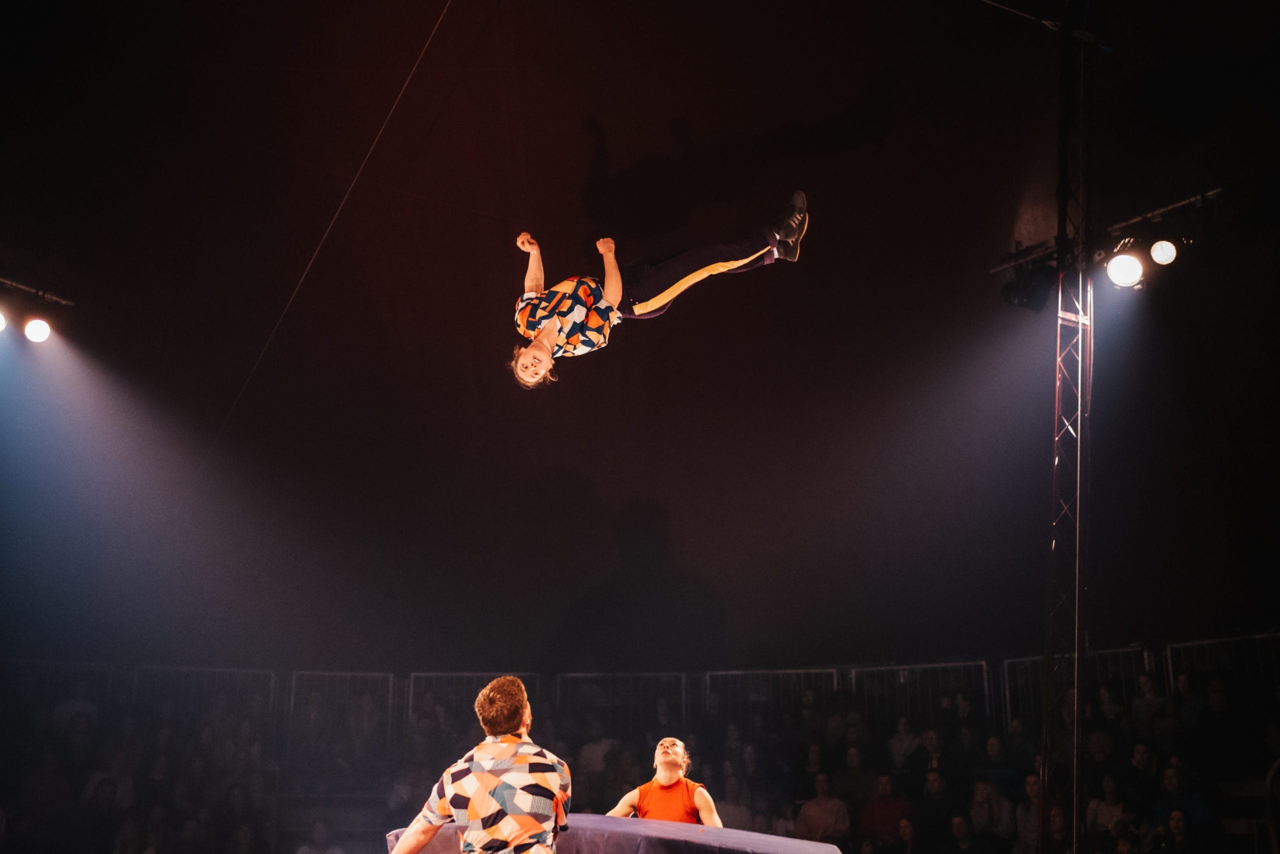 A Revel Puck circus performer is laying, parallel, in mid air. Two other performers have outstretched hands below, ready to catch their troupe-mate. Dynamic stage lights illuminate the scene.
