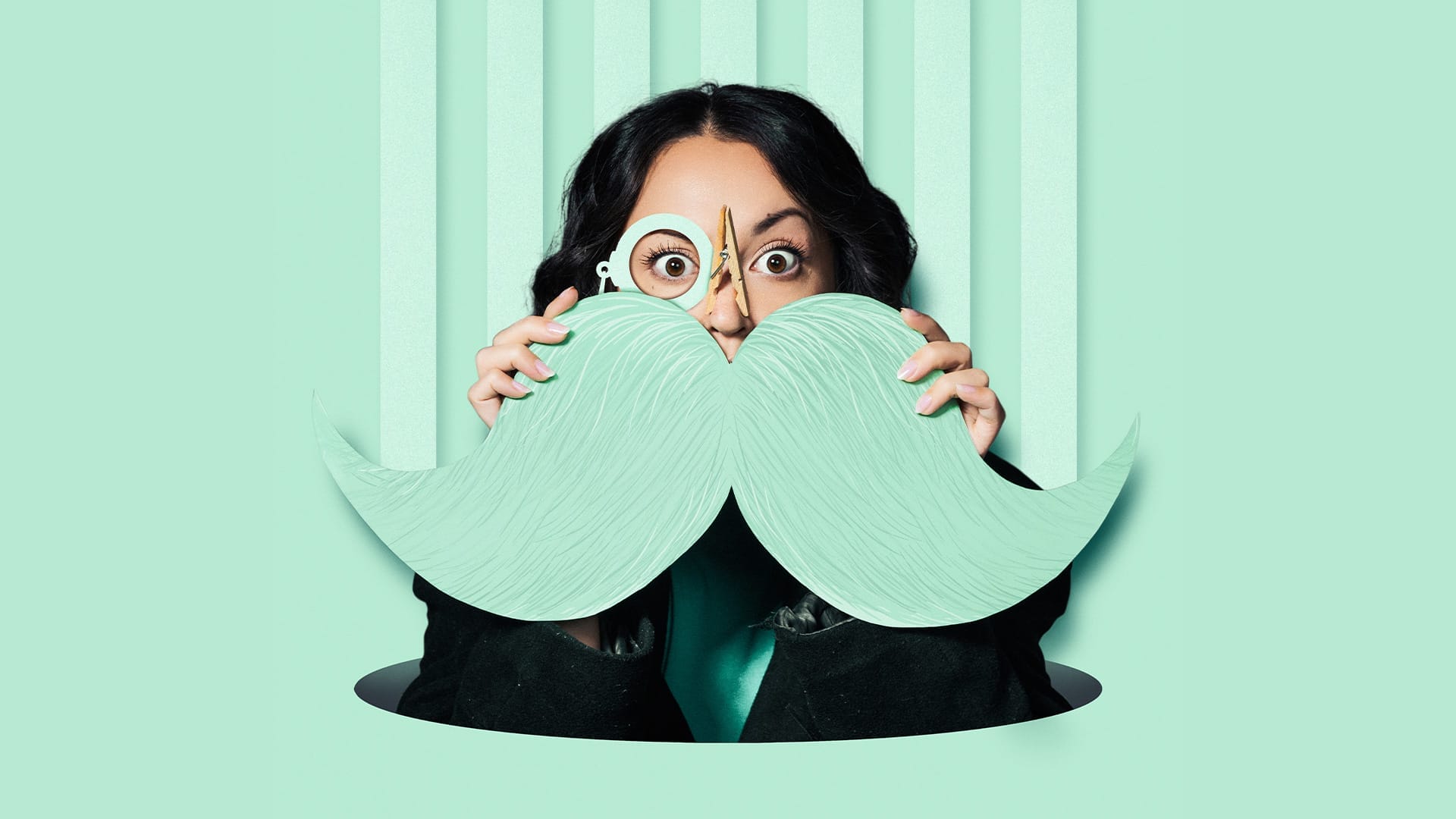 A vibrant mint green background. A woman with dark hair is holding up a large mint green cut-out moustache and monocle. Her nose is clipped with a wooden peg.