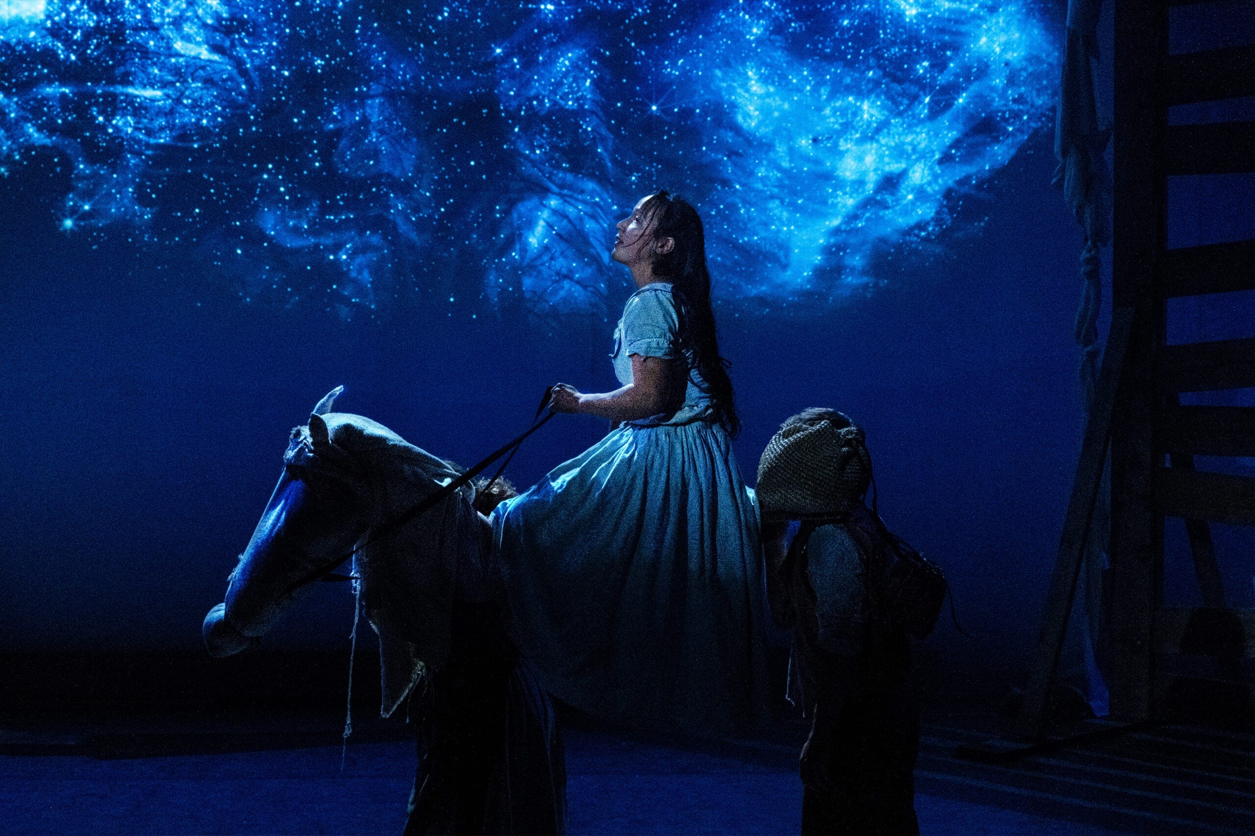 Tess is riding a horse -built from cloths and stage props. She is looking up at a mesmerising starry sky projection; dynamic blue lighting illuminates her face. She has long dark hair which falls down her back.