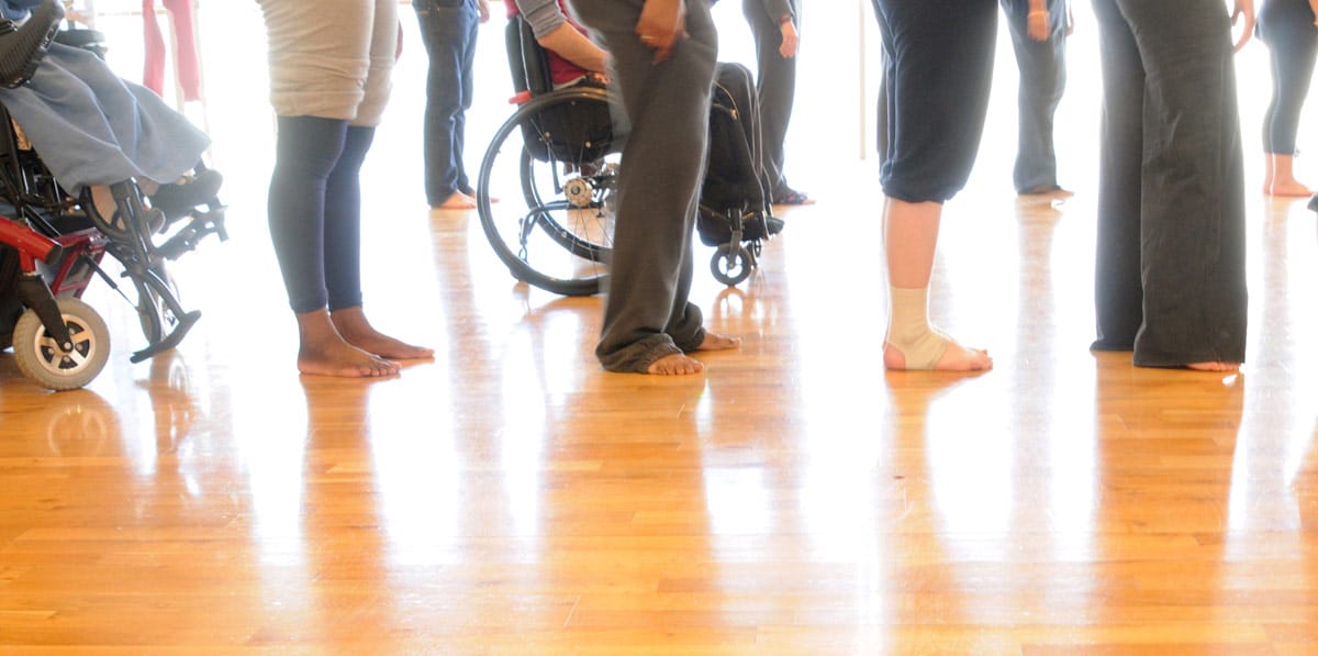 A group of people pictured, their bottom halves visible, pictured in a workshop space with a wooden floor. The group includes two wheelchair users.