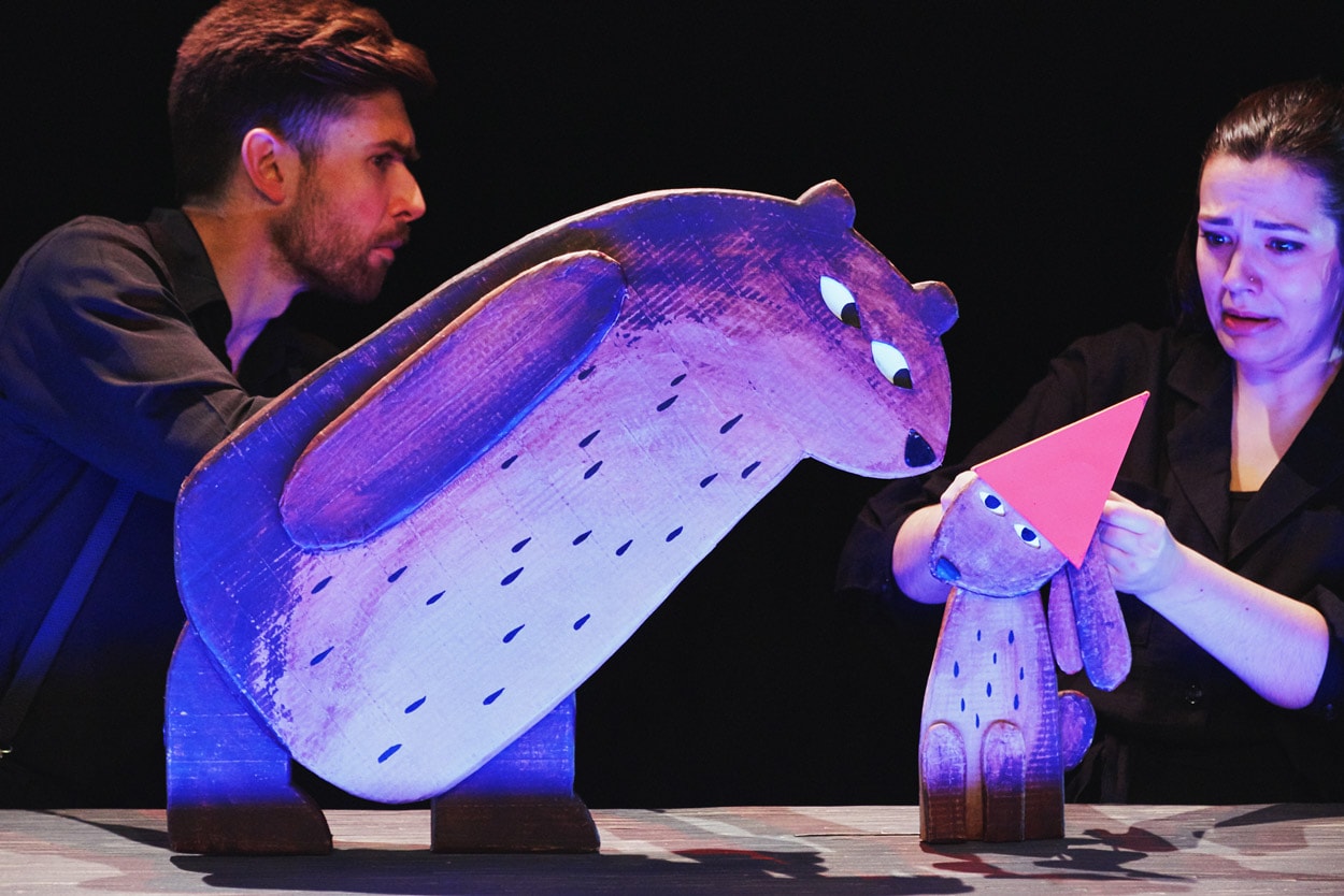 2 performers on stage in I Want My Hat Back Trilogy holding cardboard puppets of a large bear and a small rabbit wearing a hat.
