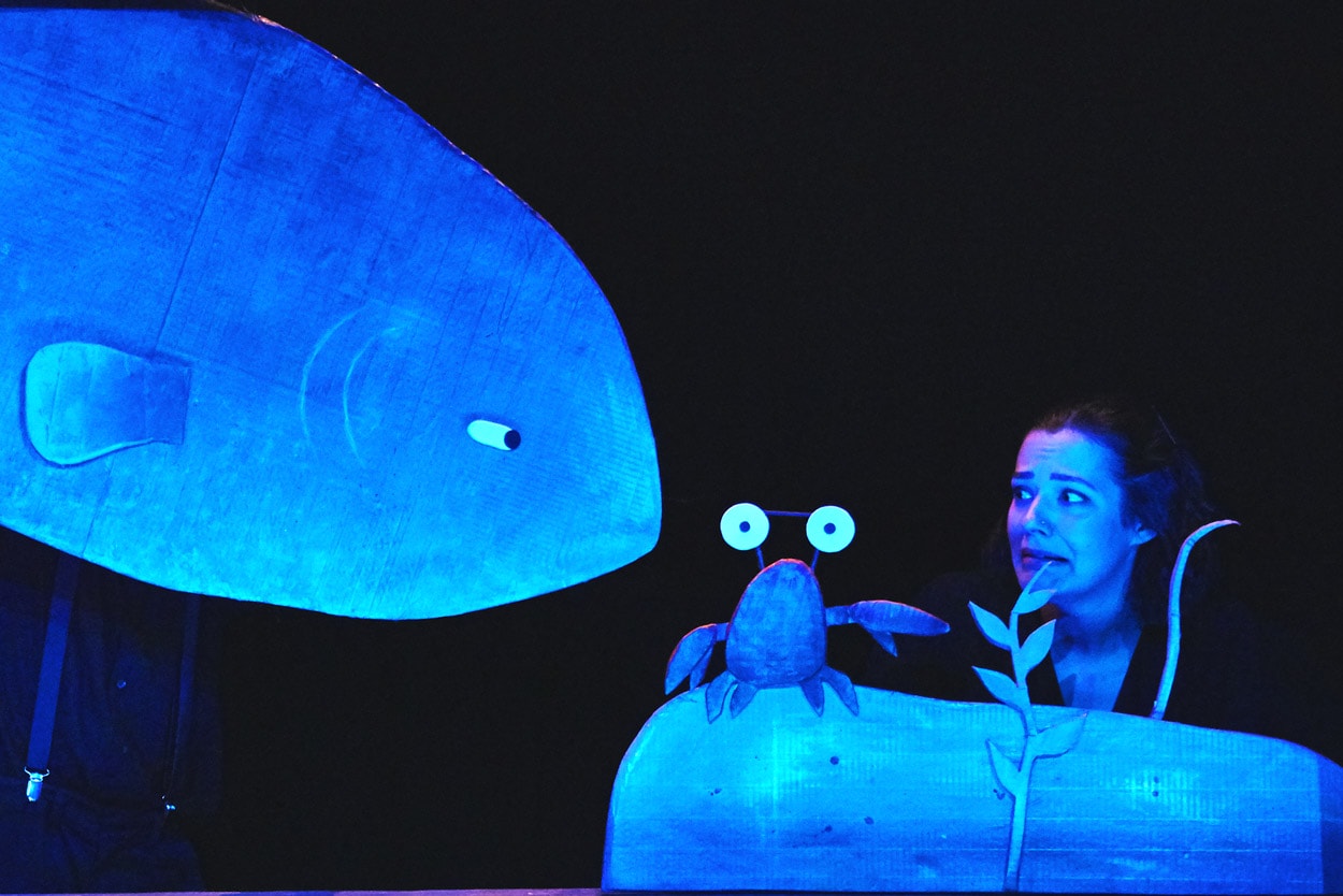 1 performer on stage in I Want My Hat Back Trilogy holding a cardboard puppet of a crab with big eyes while a large cardboard fish puppet swims into view on the left of the image