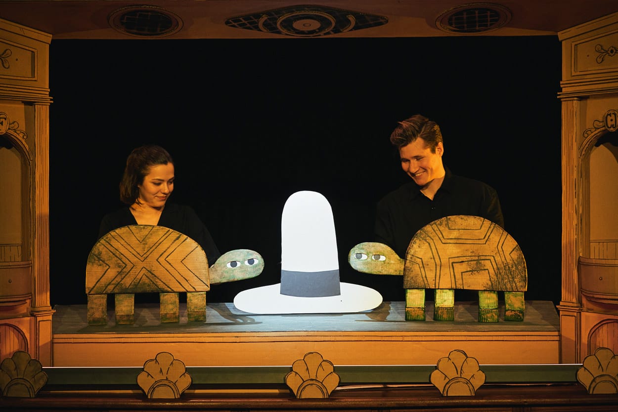 2 performers on stage in I Want My Hat Back Trilogy holding cardboard puppets of turtles. Between them is a large cardboard hat.