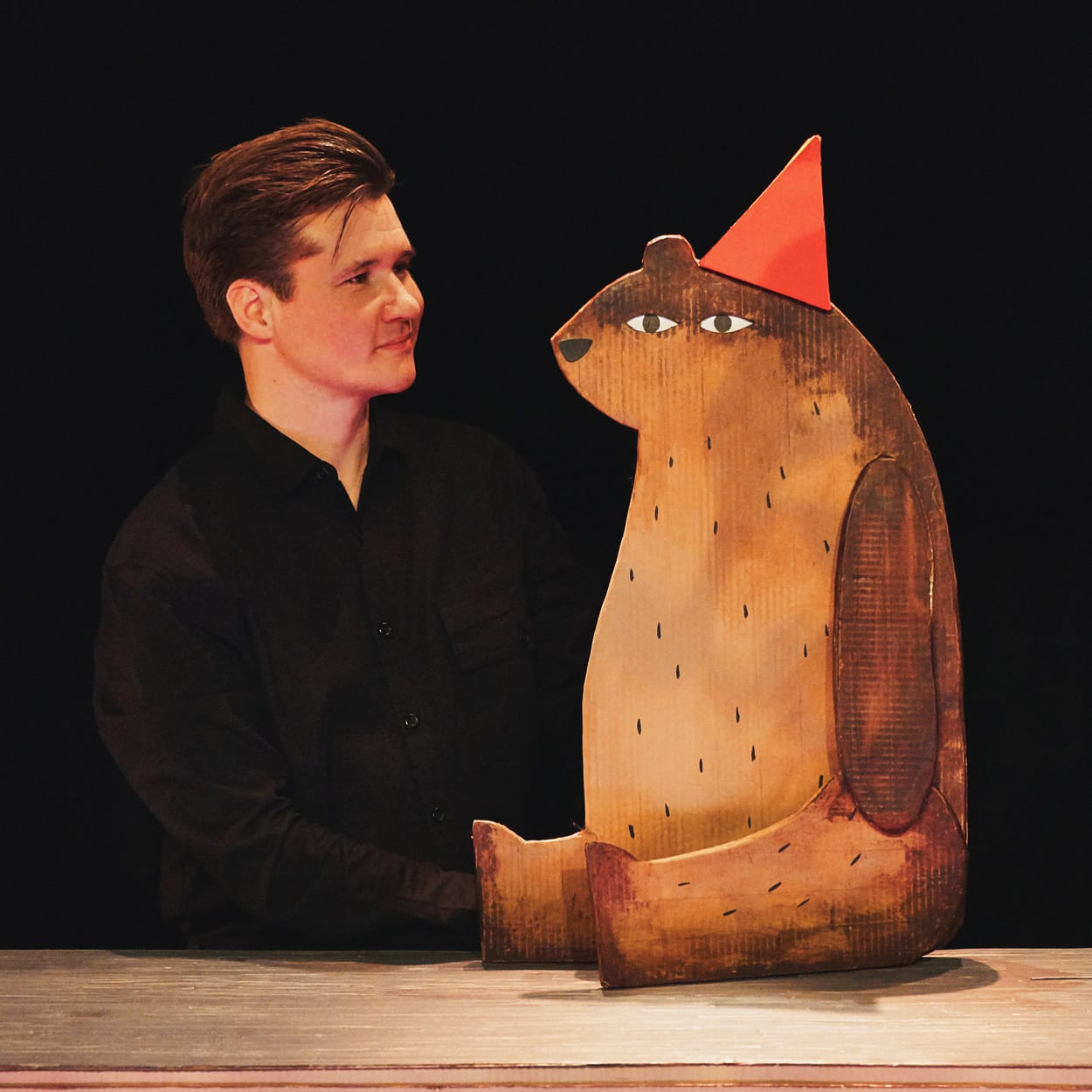 A performer on stage in I Want My Hat Back Trilogy. He is wearing black and holding a cardboard puppet of a bear wearing a hat.