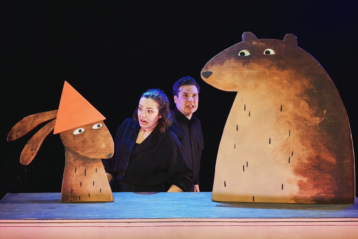 2 performers on stage in I Want My Hat Back Trilogy holding cardboard puppets of a rabbit wearing a hat and a bear without a hat.