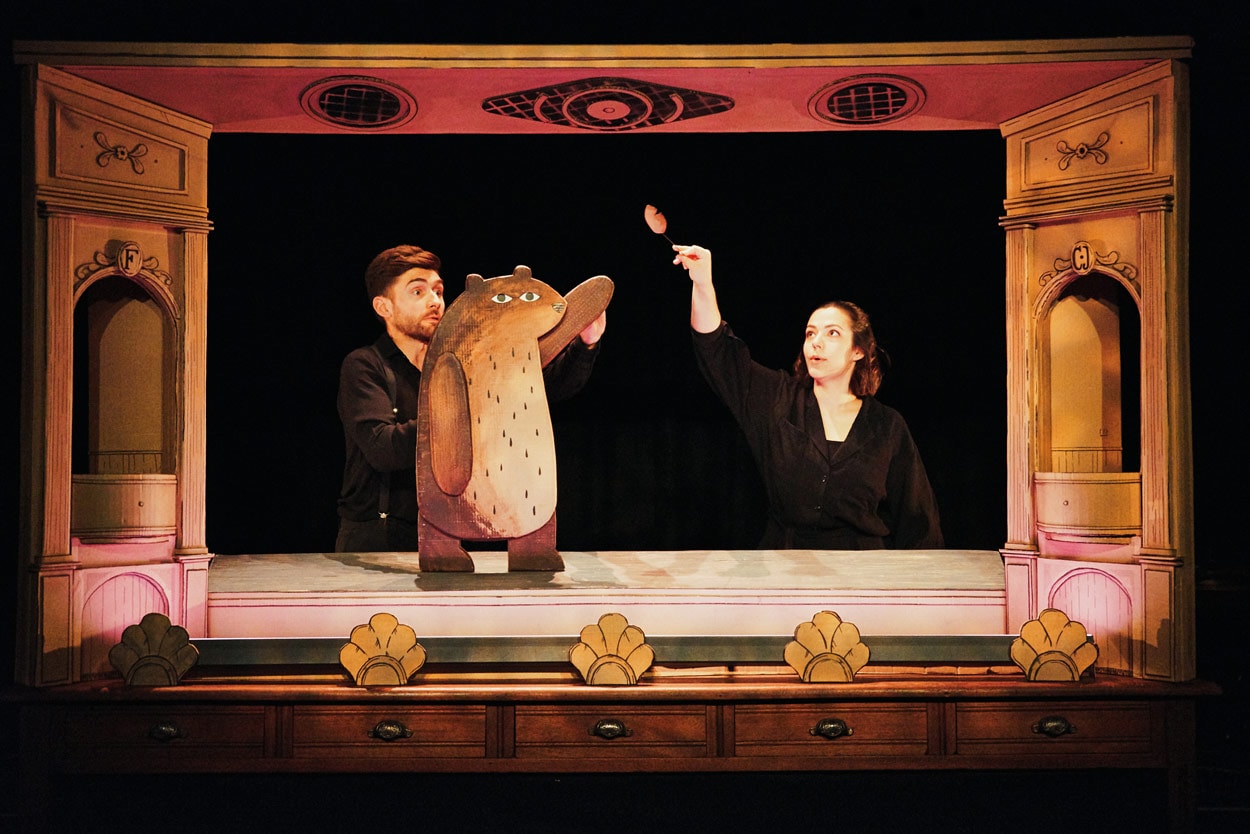 2 performers on stage in I Want My Hat Back Trilogy. They are surrounded by a cardboard proscenium arch and holding cardboard puppets - a bear and a falling leaf