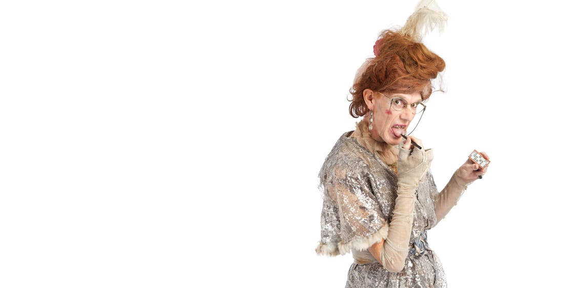 Drag performer Ida Barr wears a vintage silver dress with bat wing sleeves and a ginger beehive wig with a feather. She is holding a bingo card and licking her pen with a comical expression.