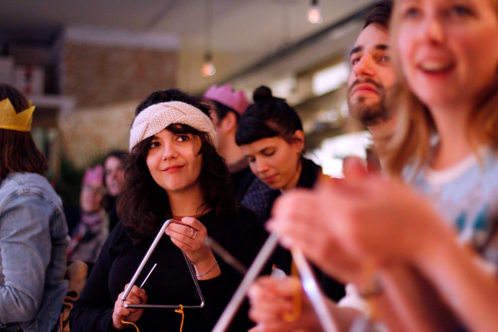 Several people standing in a crowd, one is clapping and two are playing triangles