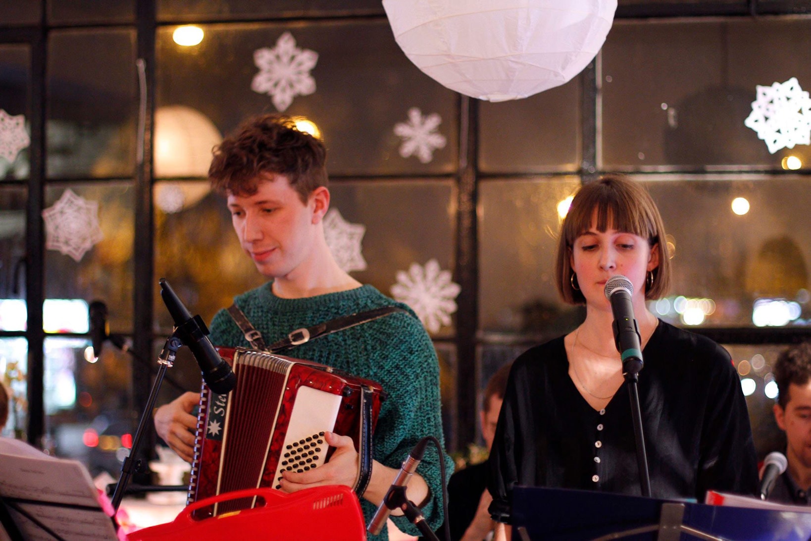 Two musicians standing behind microphones. One is wearing an accordian. Behind them cut out snowflakes are attached to a window.