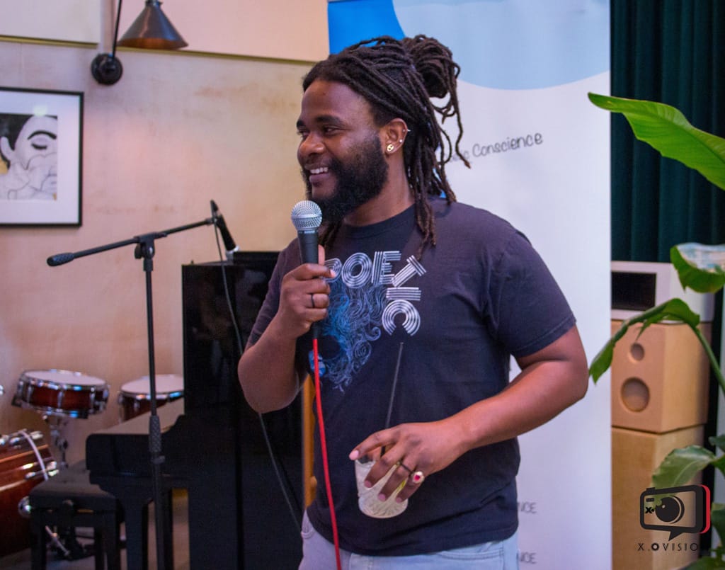 A poet performing at Poetivity. He is a smiling man with dark hair and a beard and wears a dark t-shirt. He is holding a microphone in one hand and a drink in the other.