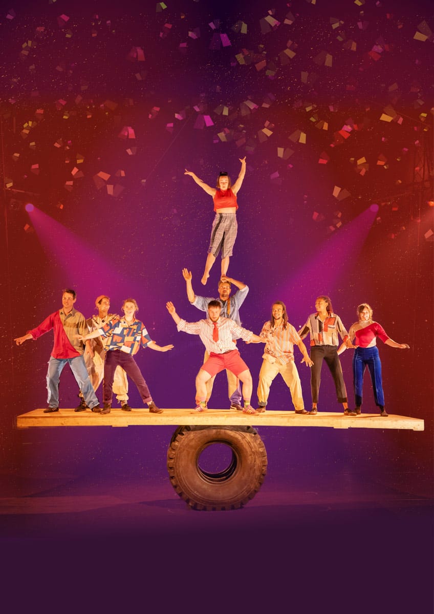 9 circus performers balancing on a plank of wood on top of a tractor tyre. One of the performers is standing on another one's head. Behind them are pink lights and confetti falls from the sky.