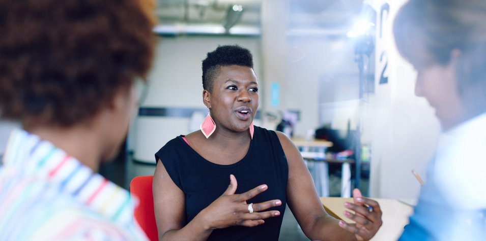 A table of three adult professionals engage in discussion at an event. In the centre, the featured speaker is a black woman with short dark hair and pink statement earrings. She is mid conversation, gesticulating with her hands.