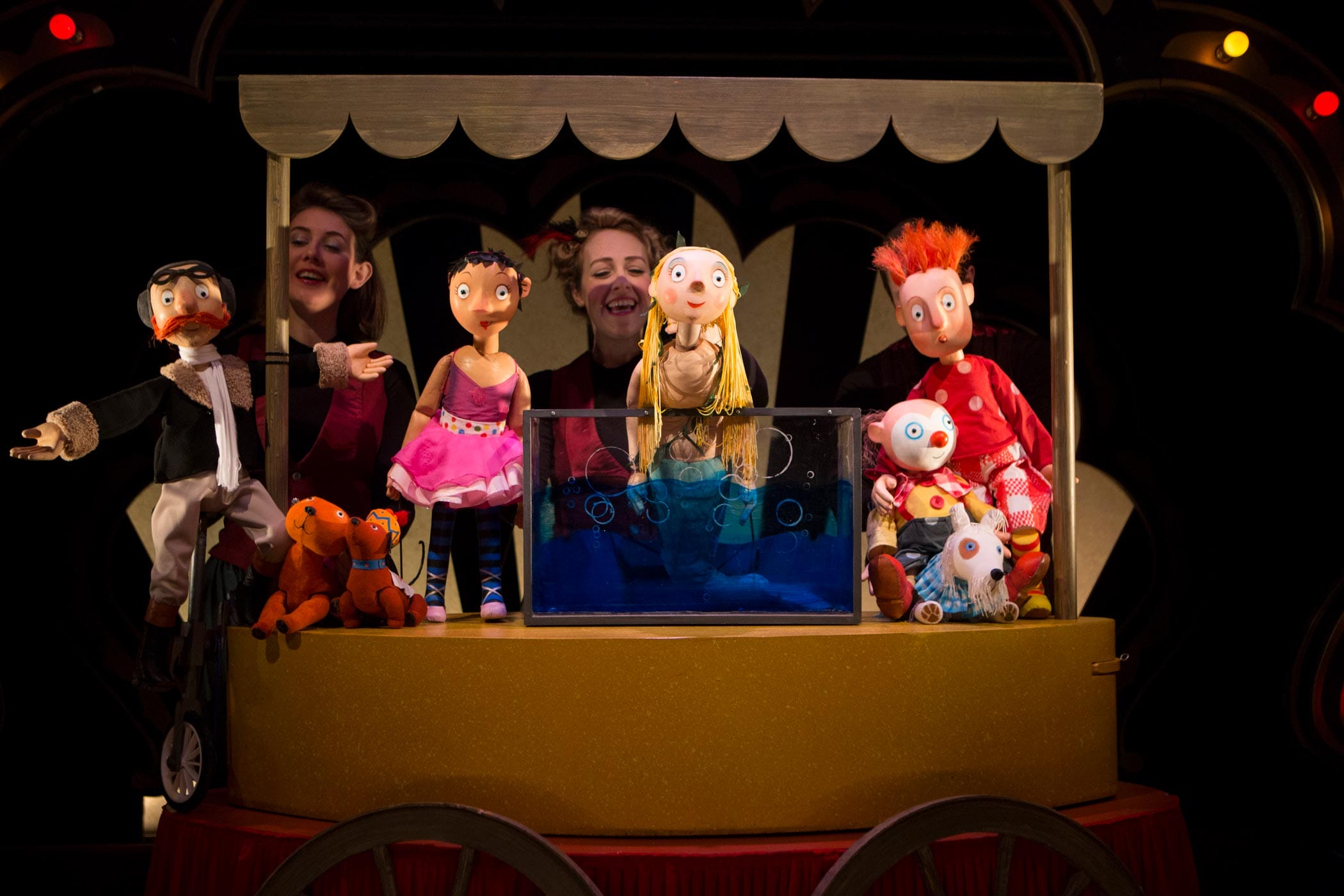 A group of circus performer puppets surround the Singing Mermaid puppet sitting happily in a tank of water. Two puppeteers can be seen in the background.
