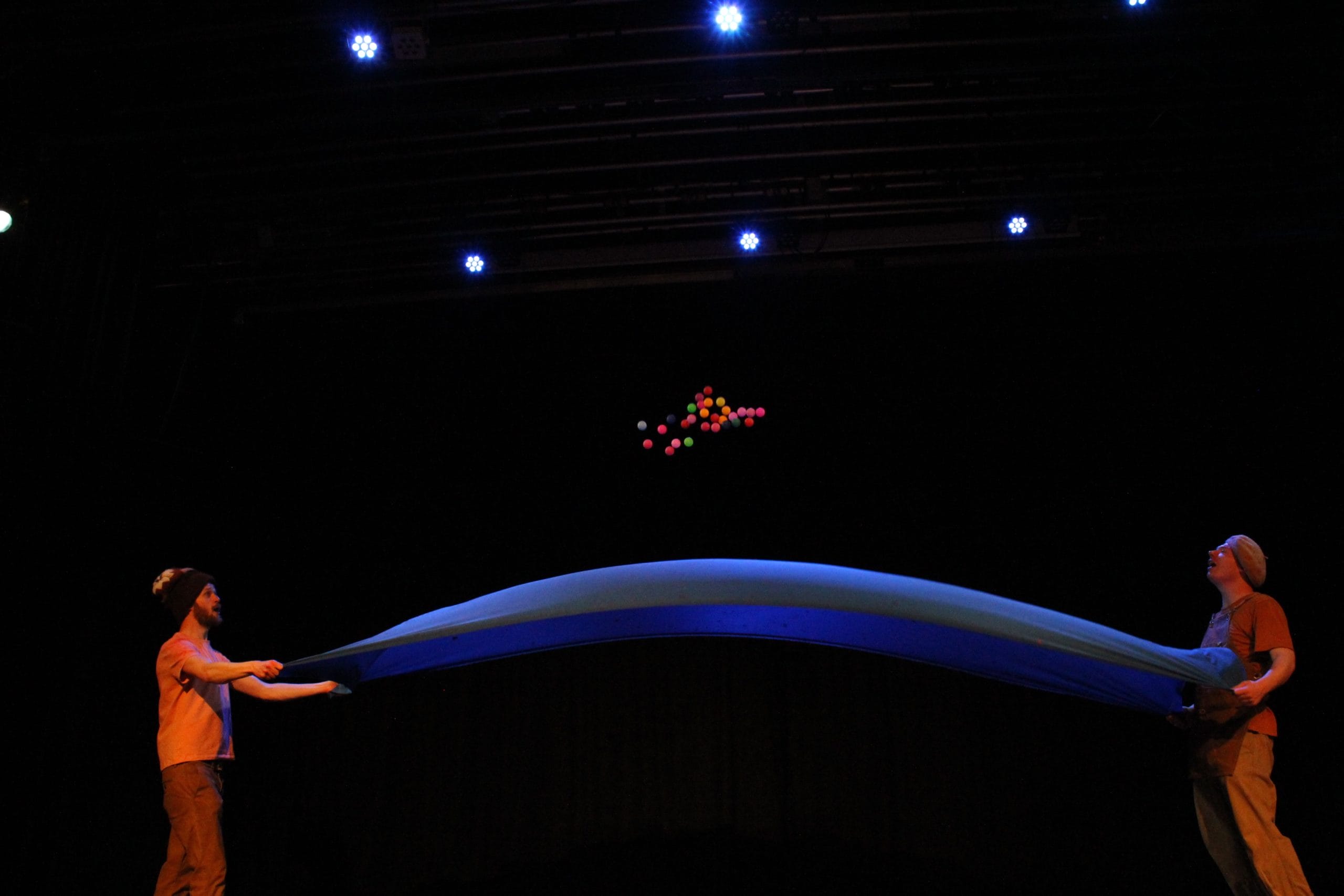Two performers stand opposite each other holding the ends of a large blue sheet. Above the sheet, they are thrusting several multicoloured balls into the air.