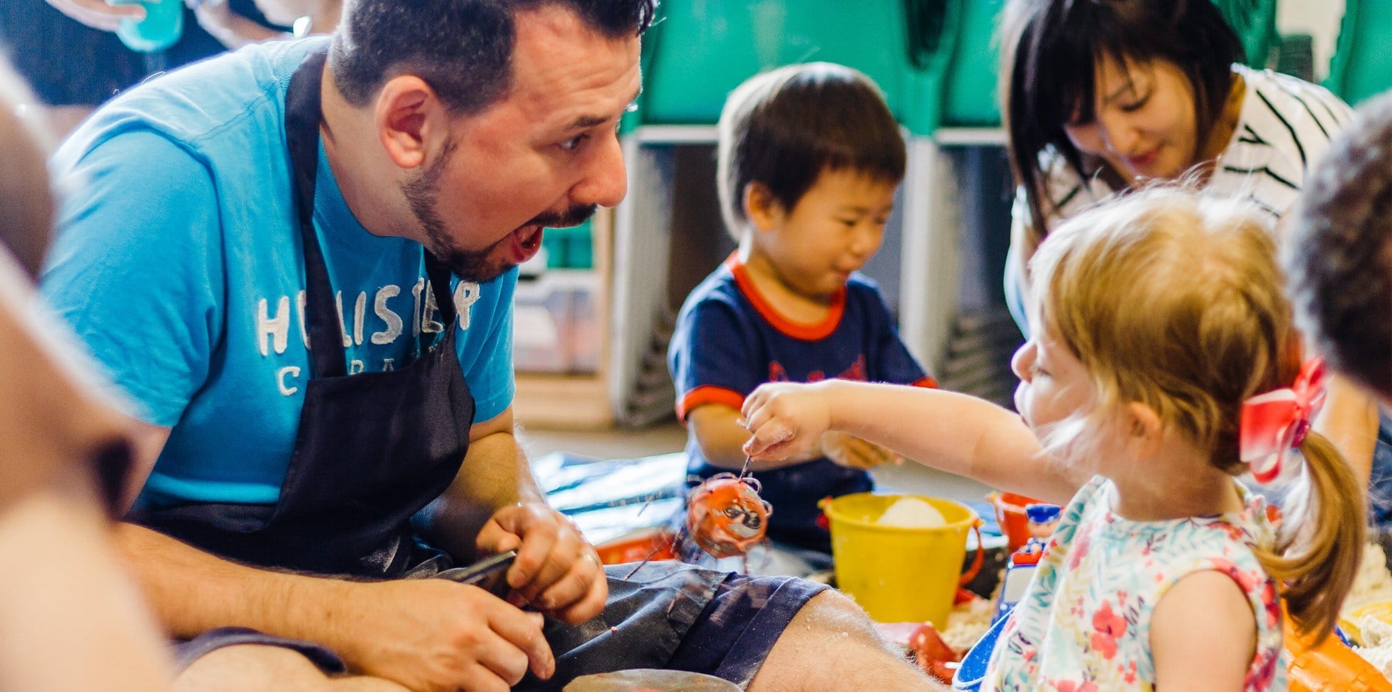 In the foreground, a smiling dad engages with his daughter as they play with a textile toy. In the background, a mum and her son are busy playing with sand and buckets.
