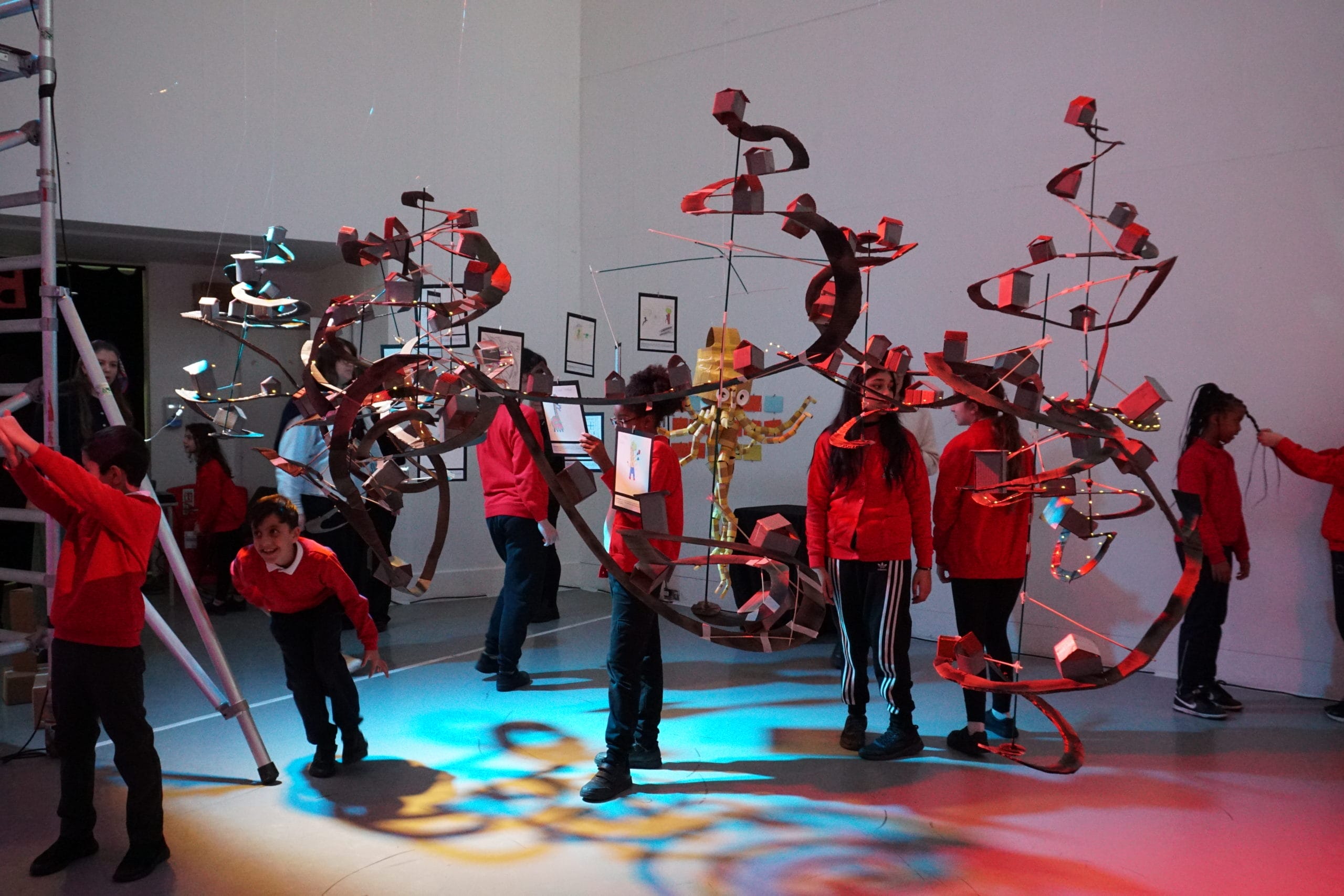 Wide shot of the exhibition space, where a futuristic world has been crafted out of cardboard and sustainable materials. Schoolchildren explore the exhibition, all wearing red uniform jumpers. Spiralling cardboard roads are hung from the ceilings with cardboard houses. The image is illuminated with atmospheric lights in blues, reds, yellows and purples.