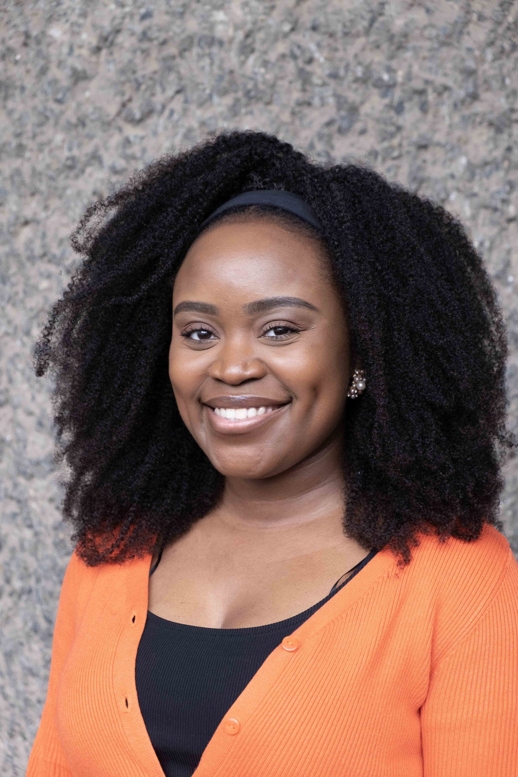 Mwansa Phiri's headshot. Mwansa is smiling and looking directly into the camera. She is a black woman with dark brown shoulder length hair and she is wearing a bright orange cardigan. Behind her is a grey pebbledash wall.