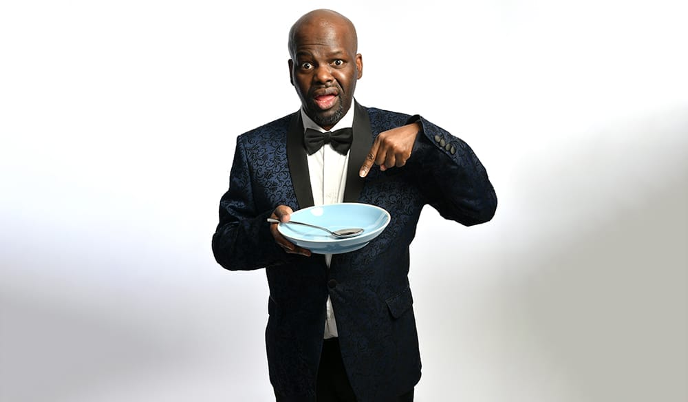Comedian Daliso Chaponda poses in a tuxedo, holding a pale blue dinner plate with a spoon. He is pointing at the bowl with a comical expression. The image background is a gray and white gradient.