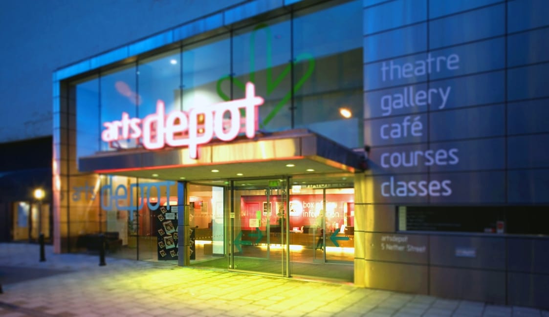 The front of artsdepot's building pictured at night with a glowing pink sign above the door and yellow light spilling out of the entrance.
