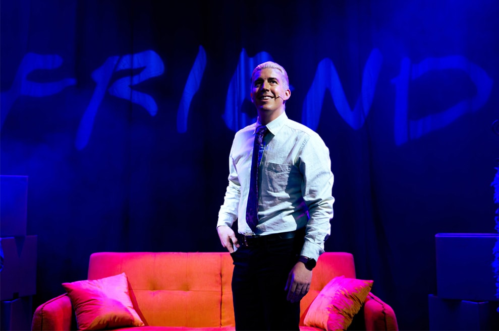 Performer stands centre stage, dressed as Gunther from friends with his signature white-blonde hair. Behind him is the signature orange couch from Friends, with the Friends logo projected behind.