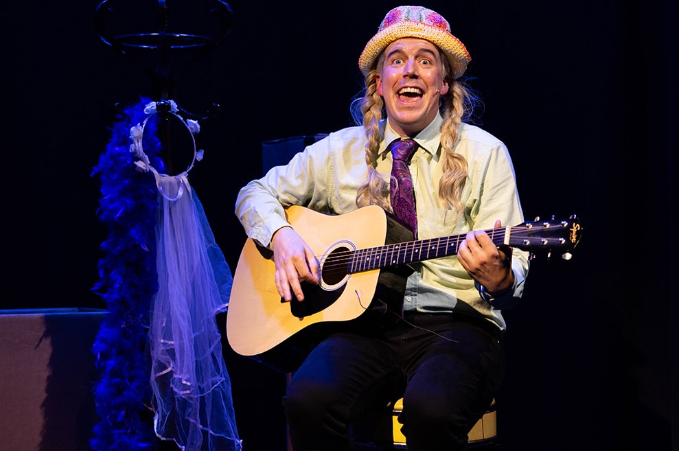 Performer is dressed as Phoebe from Friends, with blonde braids, holding her signature guitar.