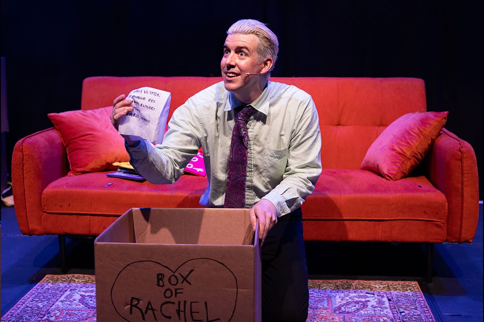 Gunther kneels in front of the signature orange 'Friends' couch. He is sifting through a cardboard box, labelled 'BOX OF RACHEL'