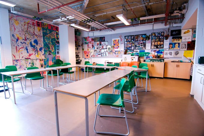 The Art Space set up for an art class. Tables are arranged in a horseshoe shape with green chairs around them. The room is full of daylight and artwork is displayed on the walls at the back of the room.