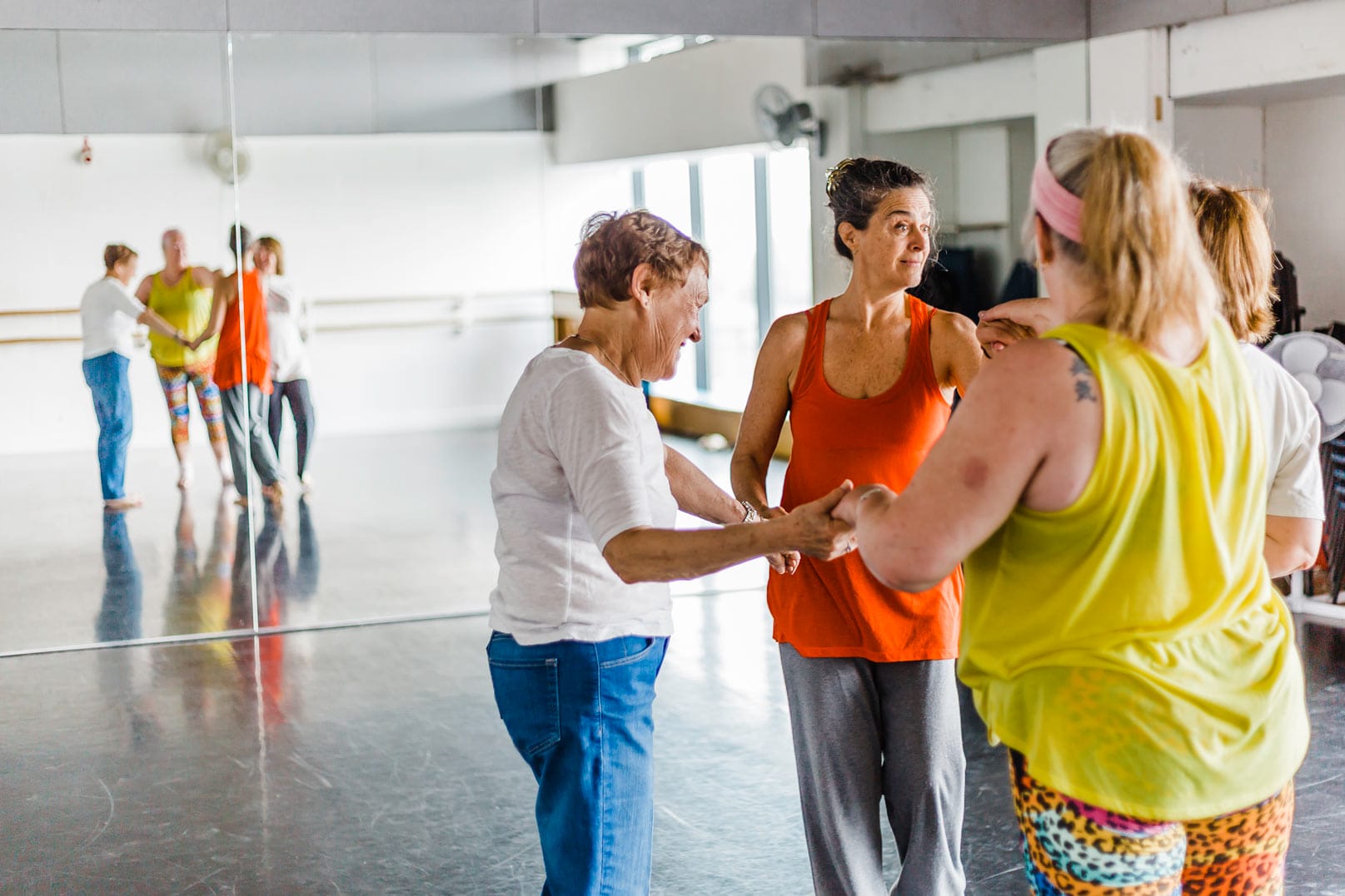 A dance taking place in the Dance Space. Four women wearing brightly coloured exercise clothes stand in a circle, their reflections can be seen in a mirrored wall behind htem.
