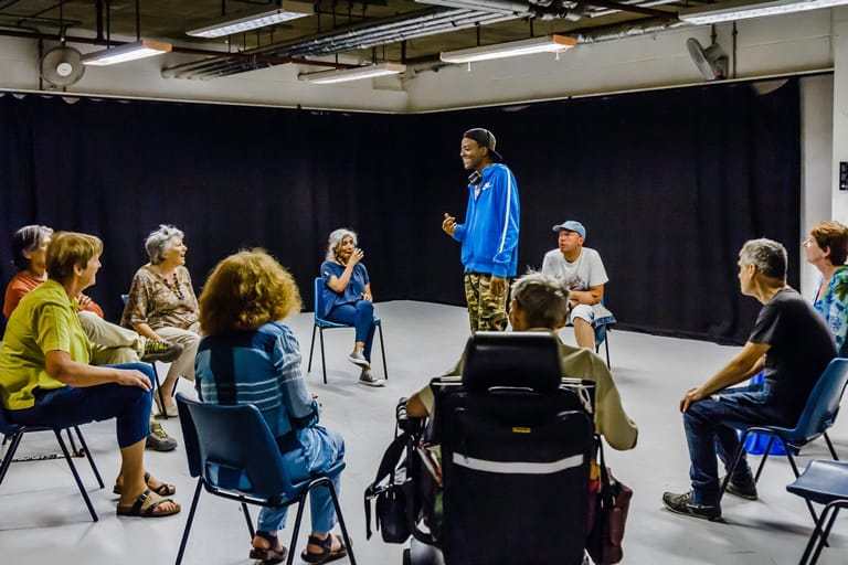 A group of people in a drama workshop in the Drama Space. A group of people are seated around a standing man. The Drama Space has a grey floor and black curtains on the walls.